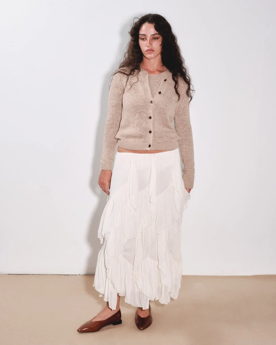 Woman wearing a beige cardigan, white layered maxi skirt, and brown heels, standing against a plain white wall.