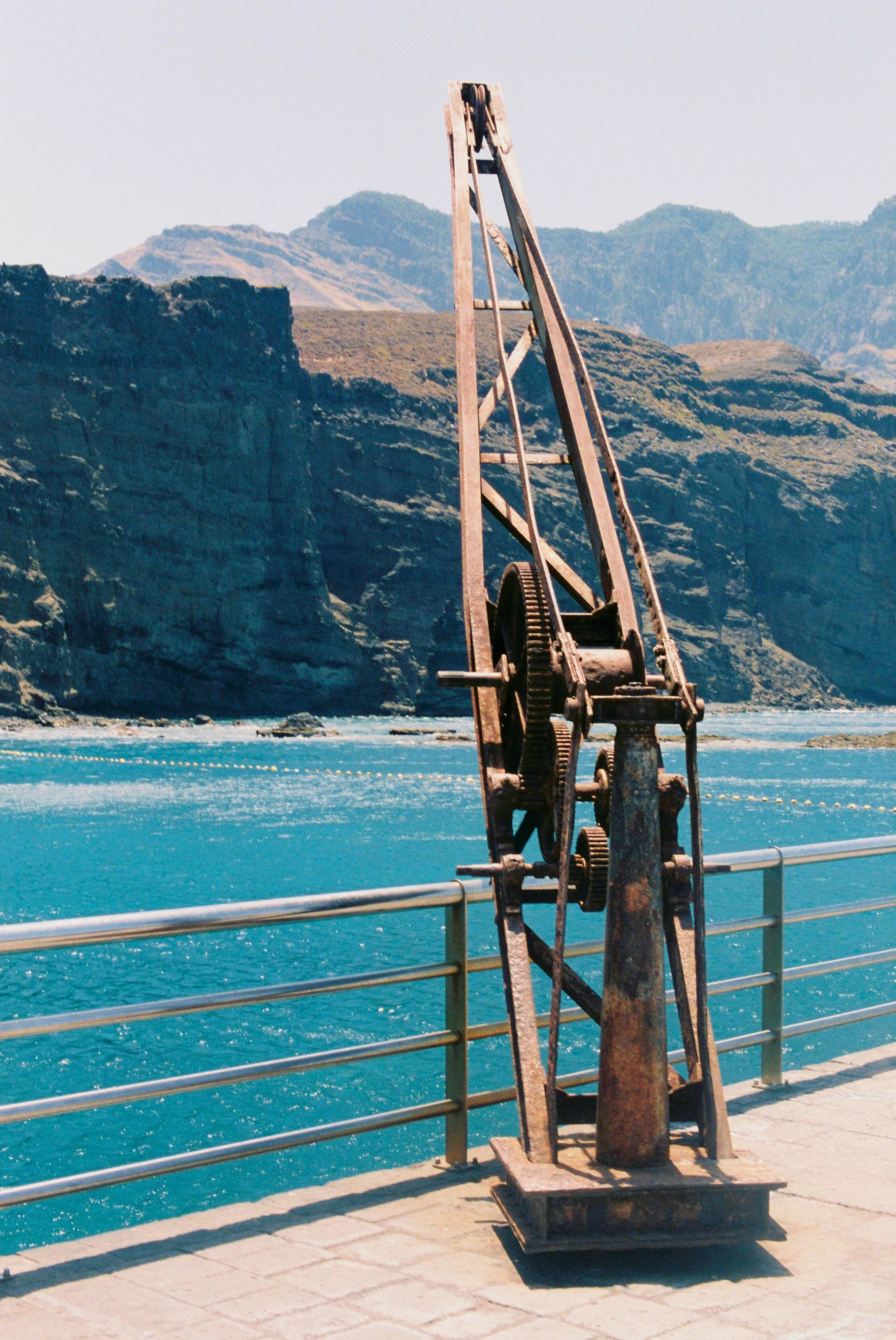 Rusty industrial gear or mechanical equipment on a dock overlooking a body of water with mountains in the background.