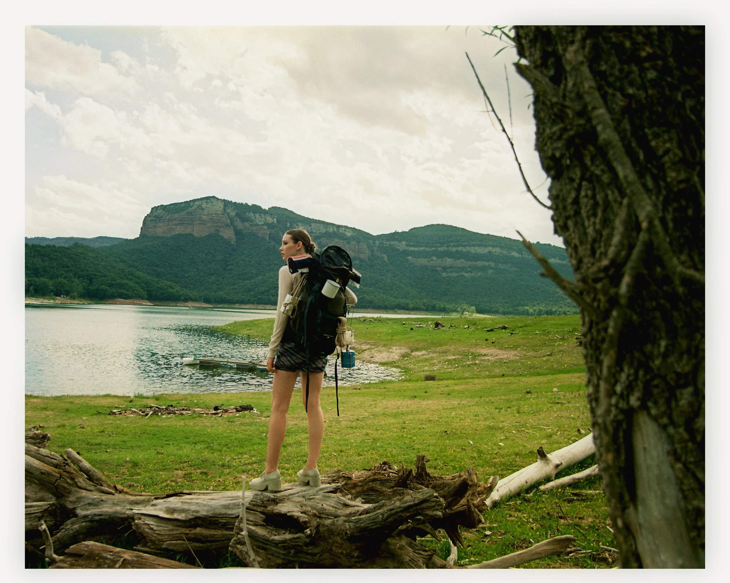A woman with a backpack standing on a fallen tree trunk near a lake, looking into the distance with mountains in the background, during daytime.