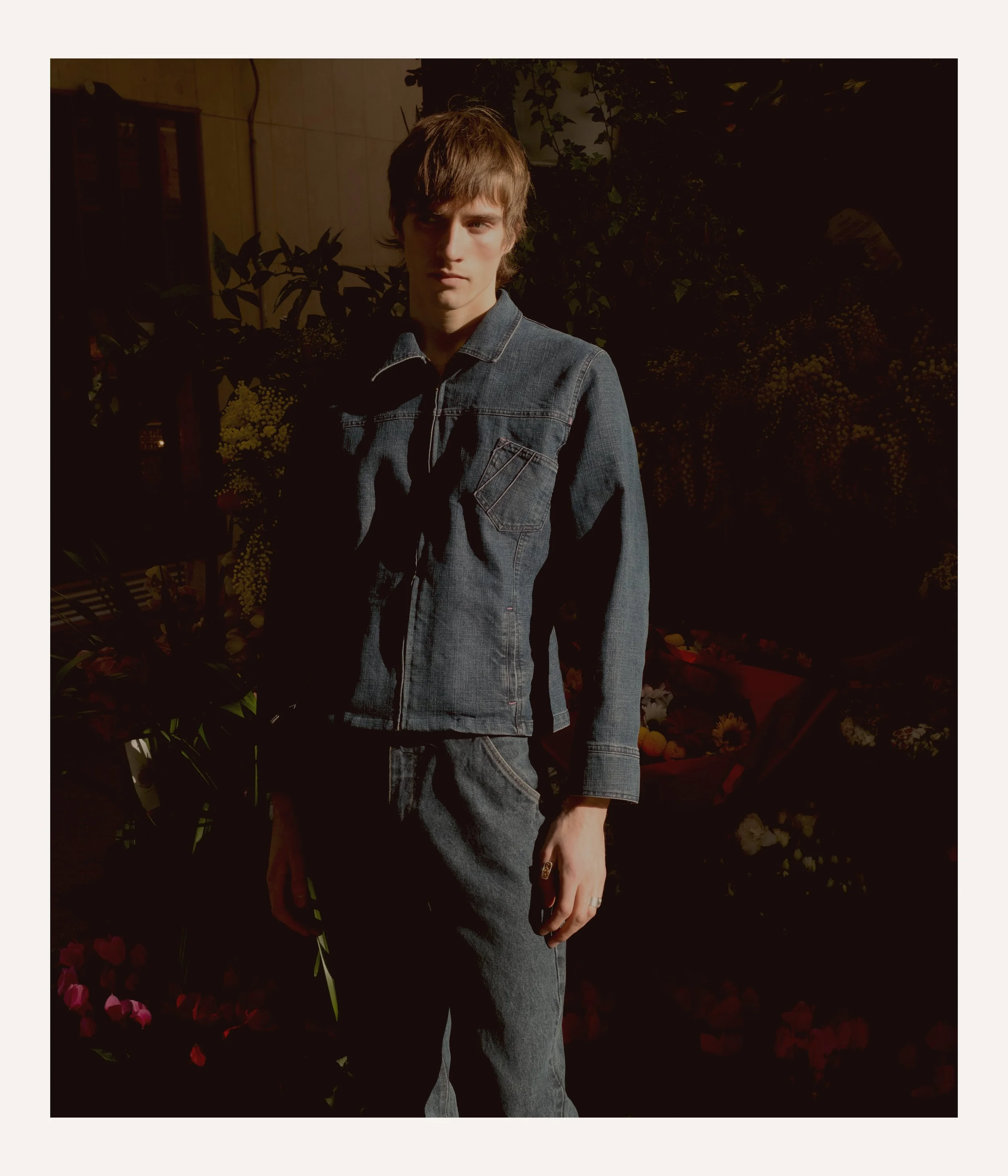 A young man with brown hair wearing a denim jacket and pants standing outdoors during daytime with plants and flowers in the background.