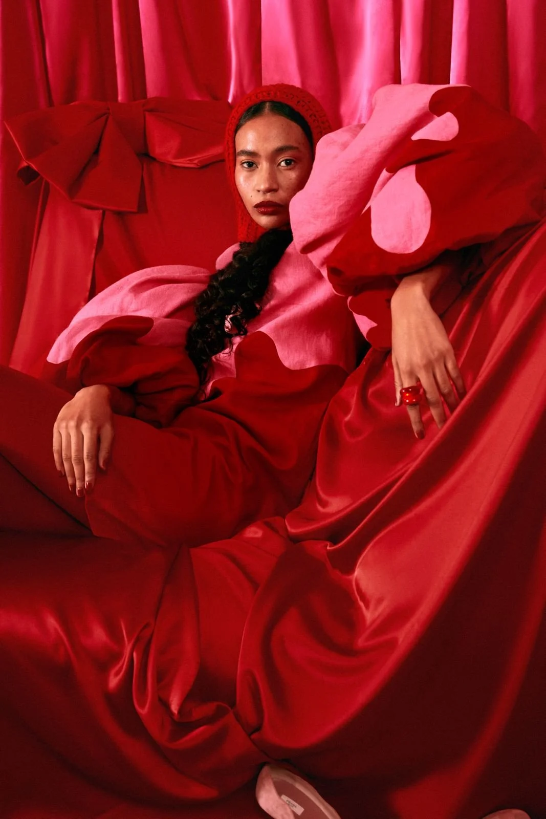 A woman in an all-red outfit, including a red knit hat, sitting on a red upholstered chair with red curtains in the background, looking directly at the camera.