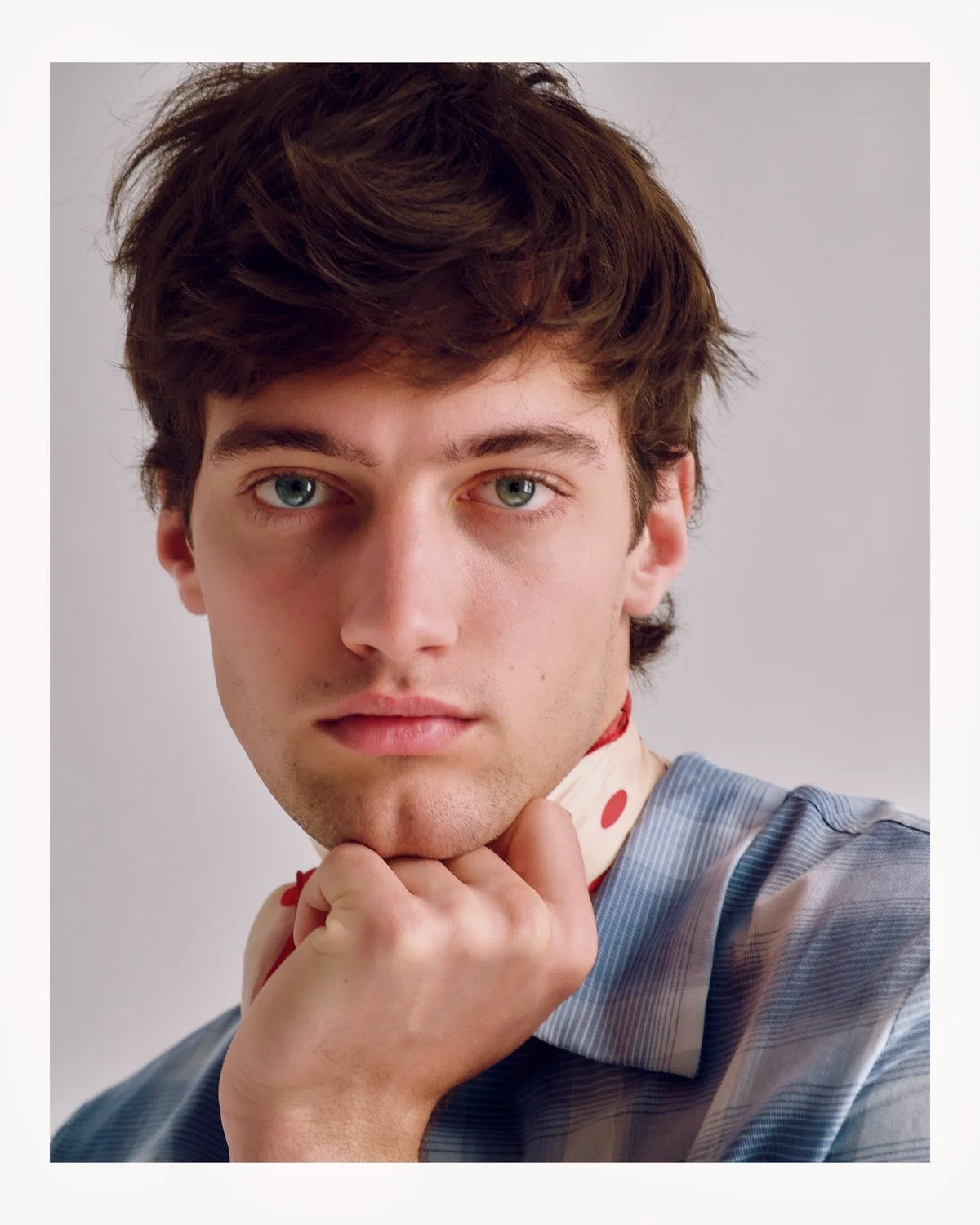 Close-up portrait of a young man with brown hair and blue eyes, resting his chin on his hand, wearing a striped shirt with a polka dot necktie.