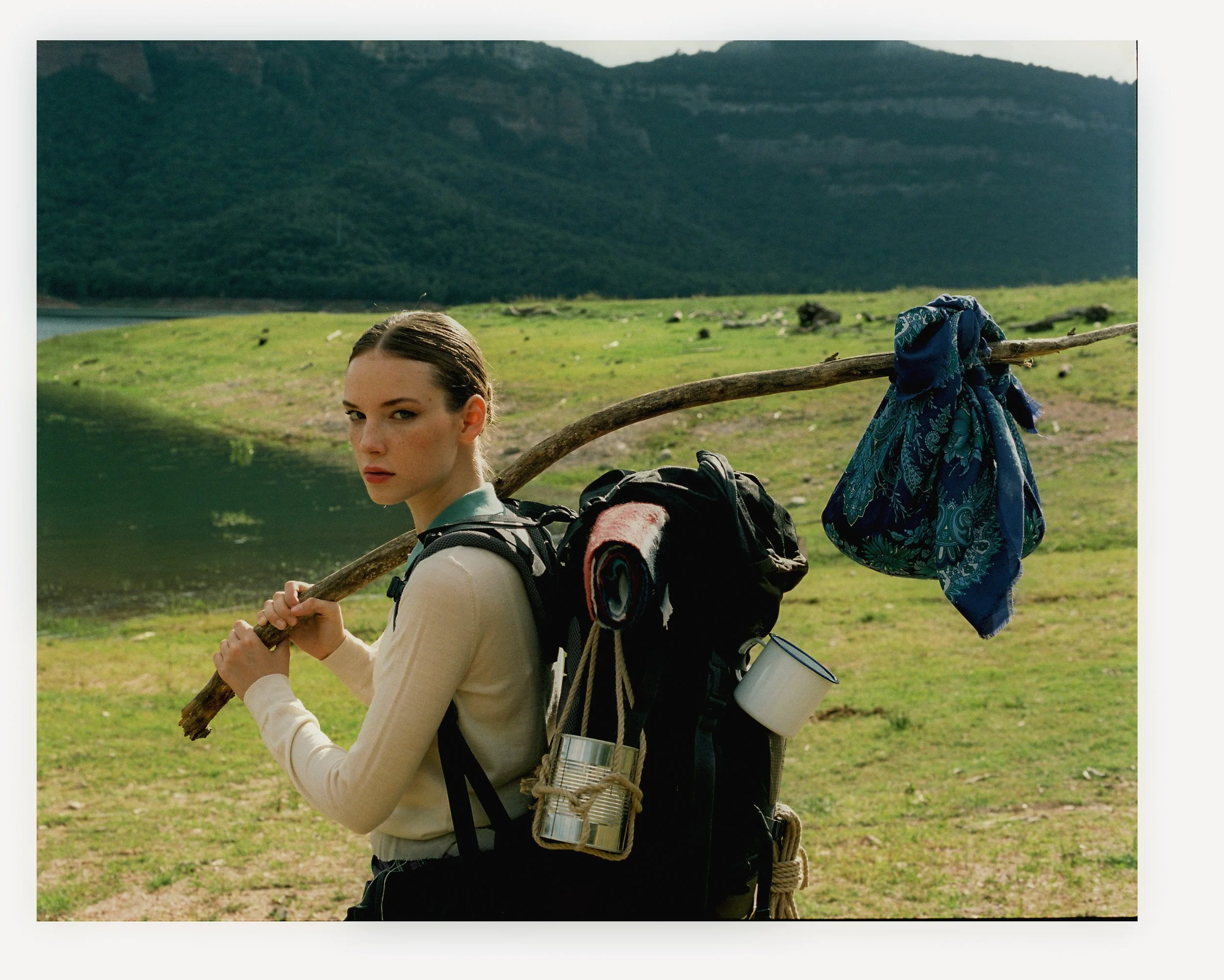 Woman with a backpack carrying a staff and a blue cloth tied to it, standing by a lake with mountains in the background.