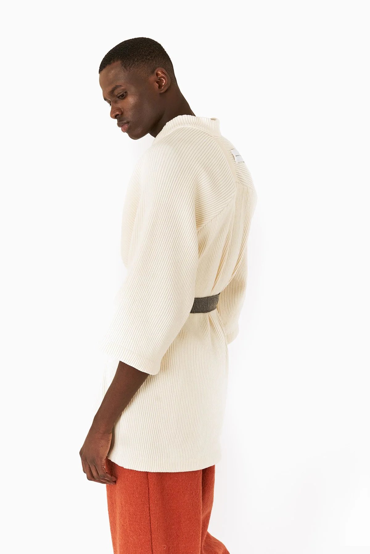 A young man with dark skin and short haircut wearing a cream ribbed knit sweater with a high neck, and burnt orange pants, standing against a white background, looking downward with his body turned slightly to the side.