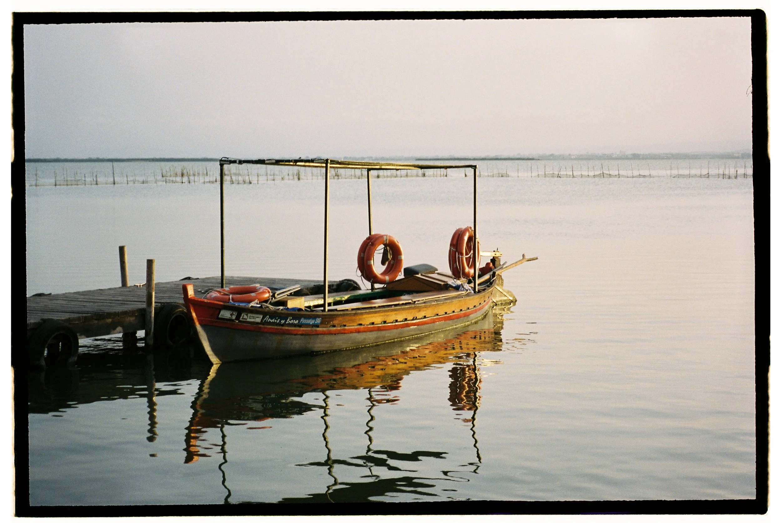 A boat docked at a small pier on calm water with life rings on board and a floating dock attached.