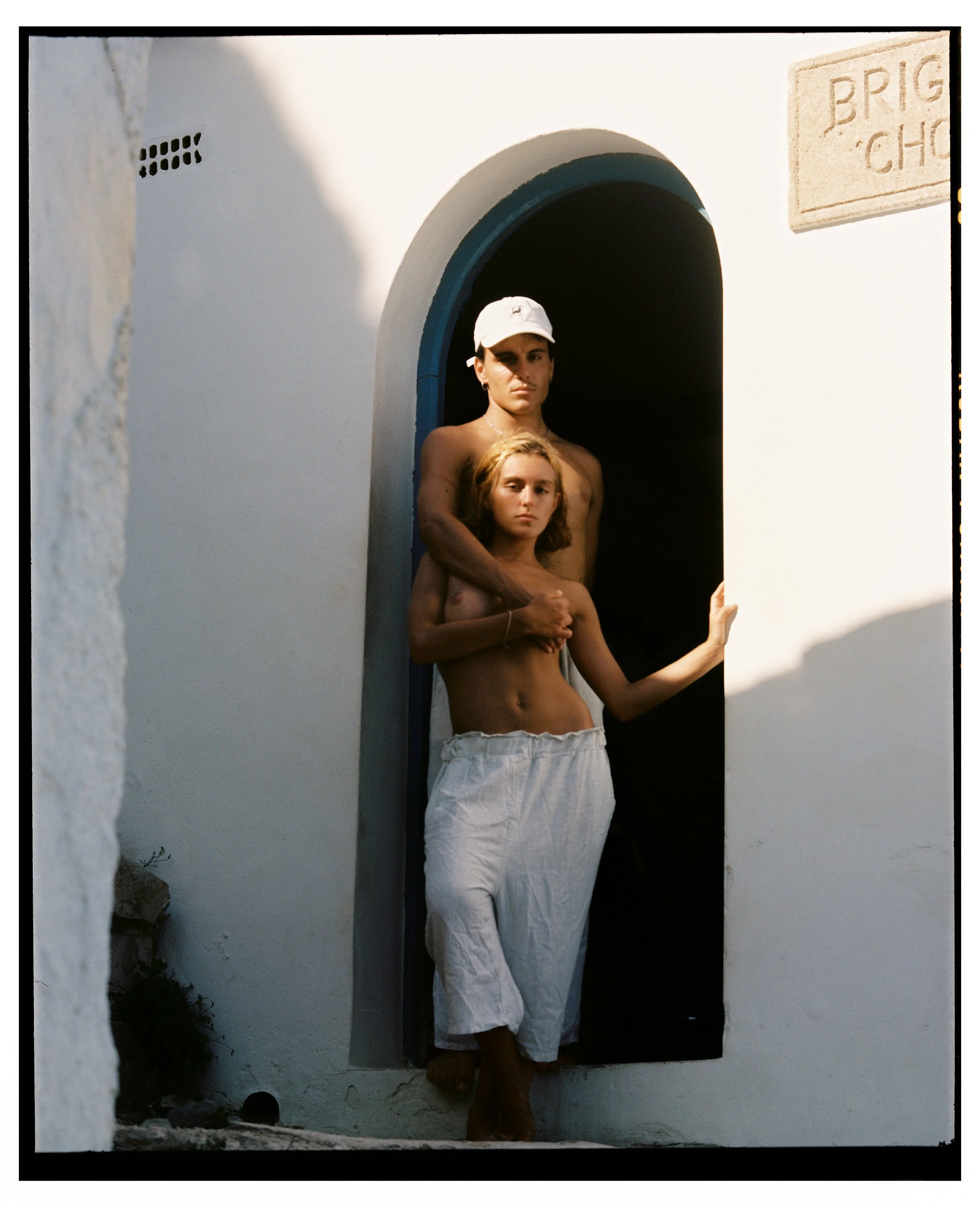 Two shirtless women stand in an arched doorway of a white building; one wears a white baseball cap and the other has shoulder-length blonde hair, both with serious expressions.