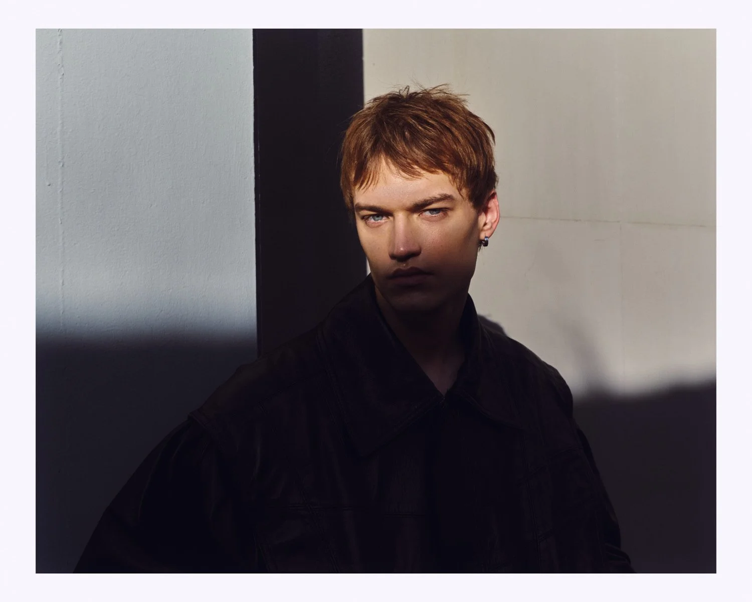 Young man with reddish hair wearing a black jacket, standing against a wall with a shadow cast across his face, in a dimly lit setting.