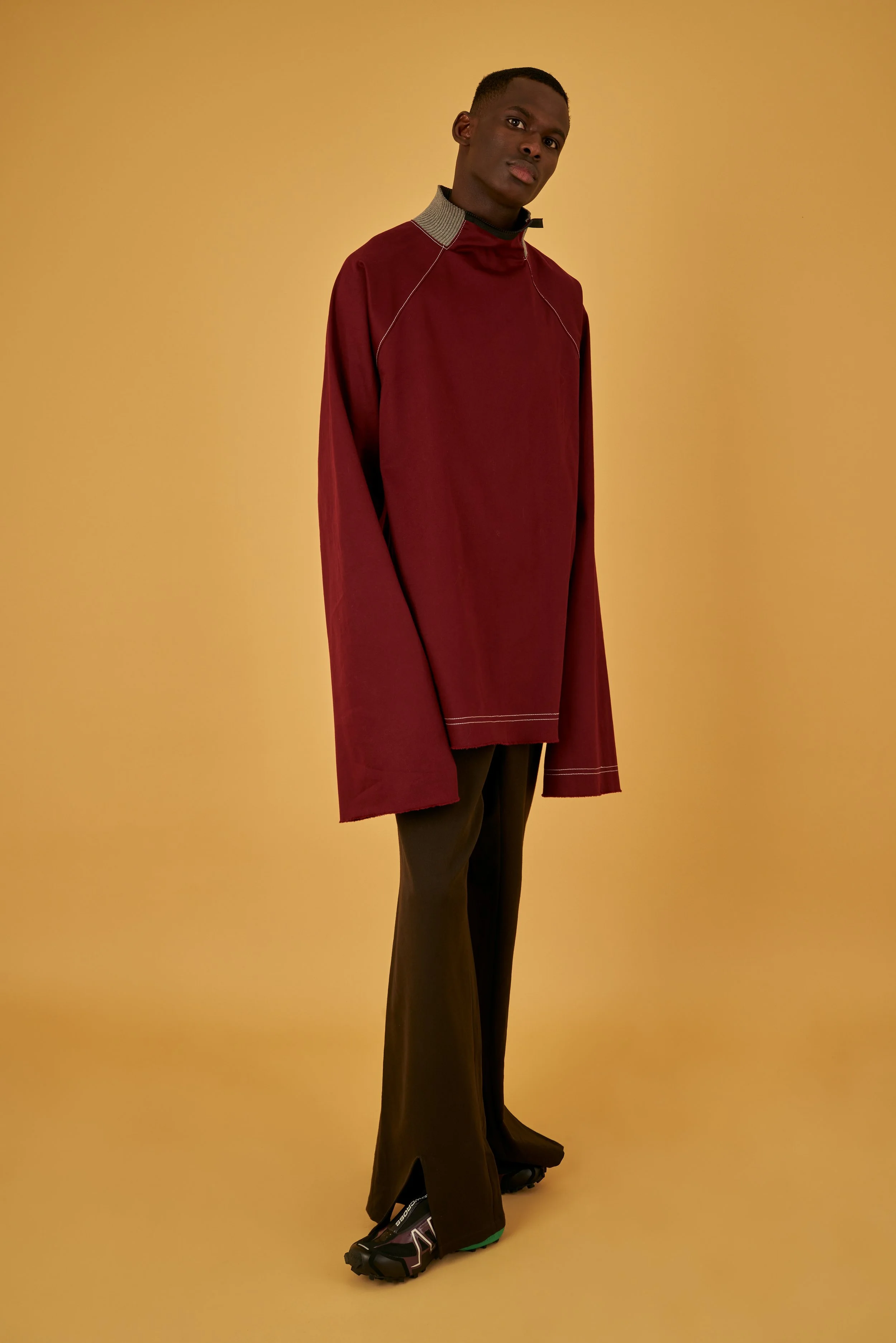 A tall man with dark skin and short hair dressed in a maroon oversized top with contrasting collar details, black pants, and running shoes standing against a plain beige background.