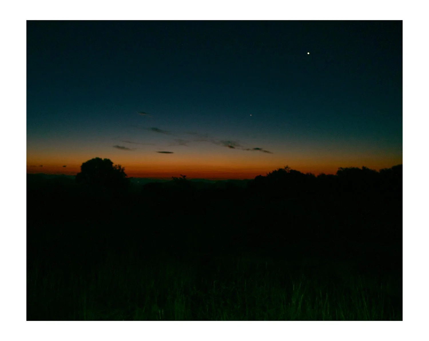 A sunset or sunrise over a rural landscape with dark silhouettes of trees and a grassy field in the foreground, with a twilight sky featuring orange near the horizon and dark blue above, with a few visible stars.