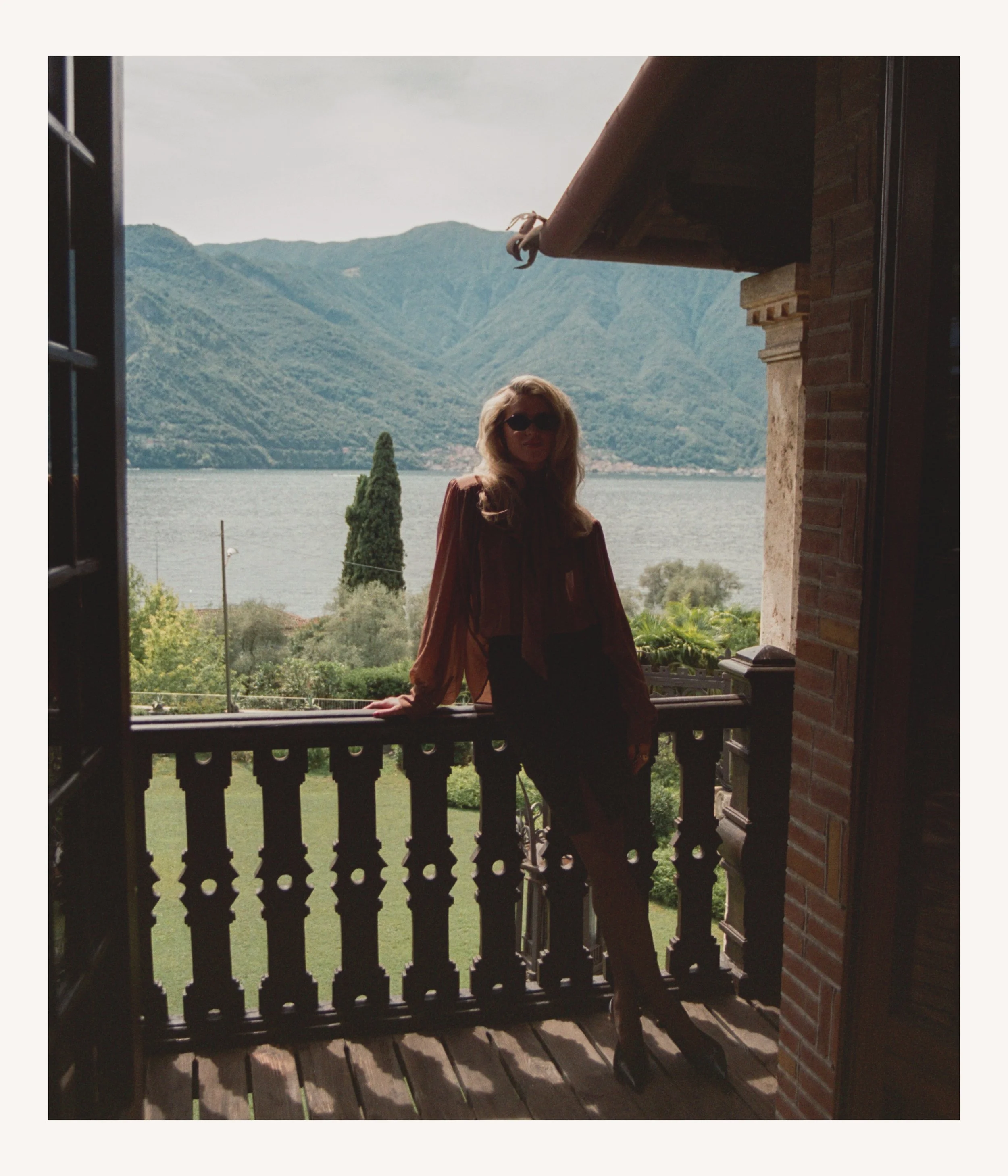 A woman with long hair and sunglasses standing on a balcony with a view of a lake and mountains in the background.