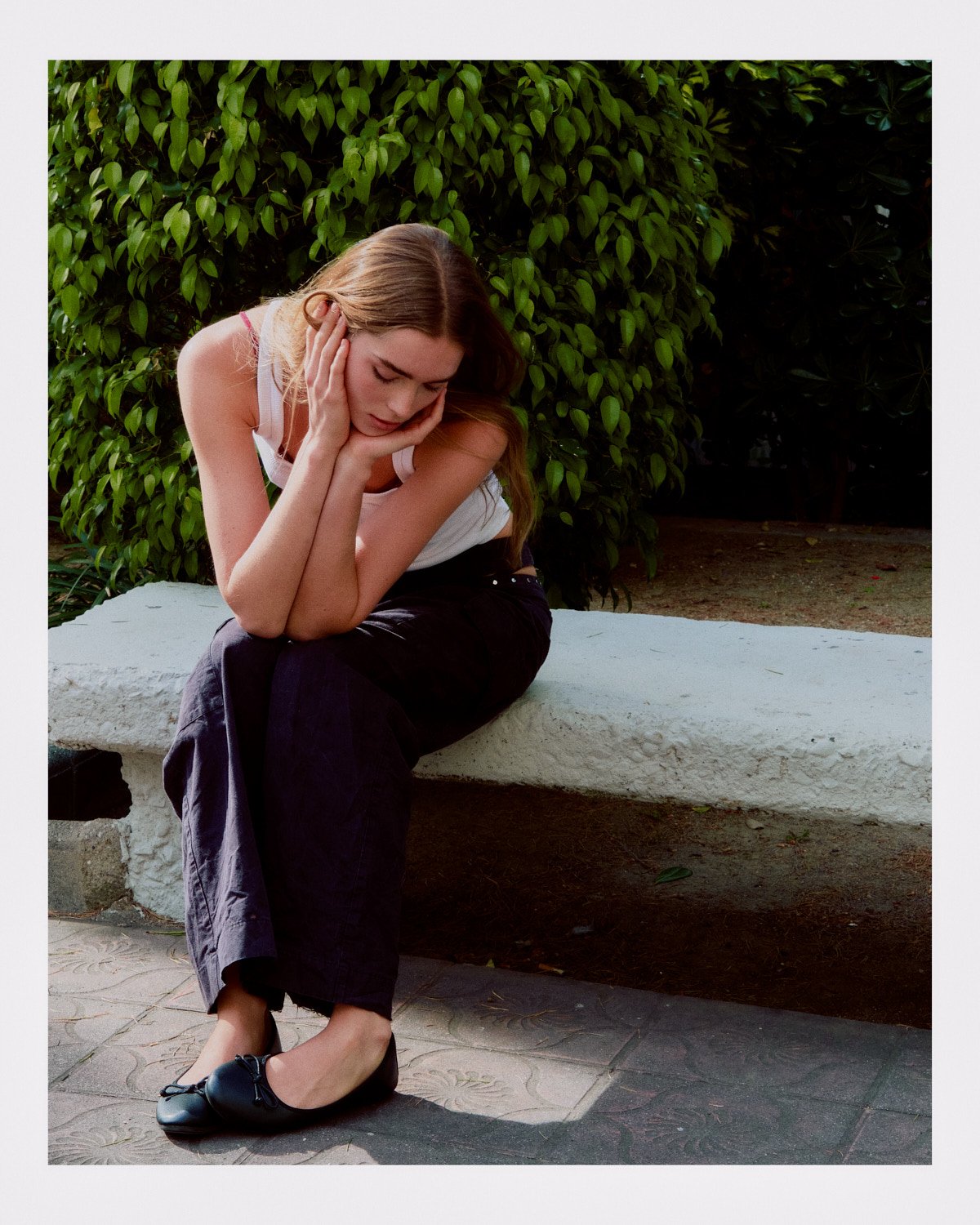 Young woman sitting on a white stone bench, looking sad, with her head resting on her hand, outdoors surrounded by greenery.