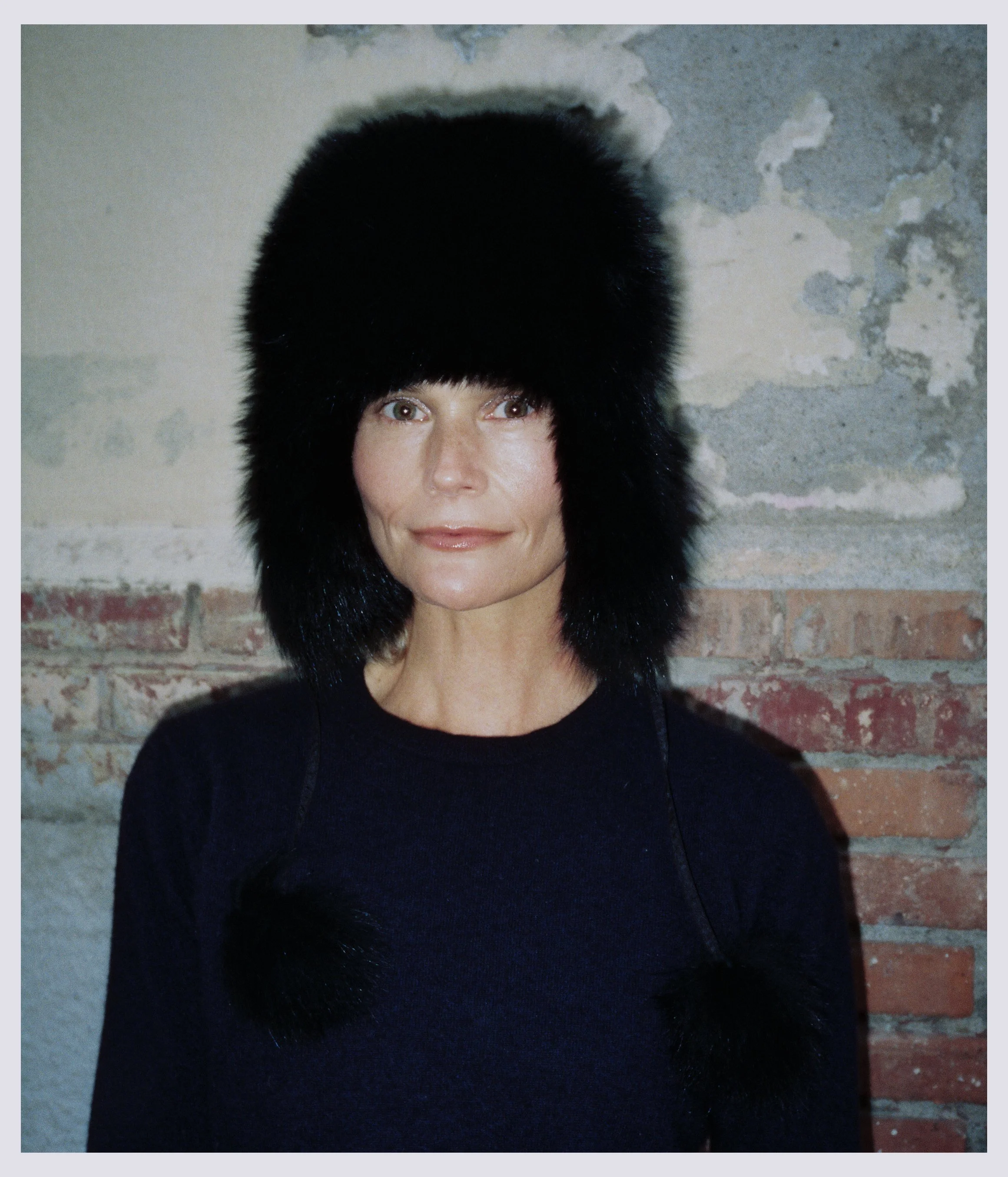 Woman wearing a large black fur hat with pom-poms, standing against a textured brick and concrete wall.