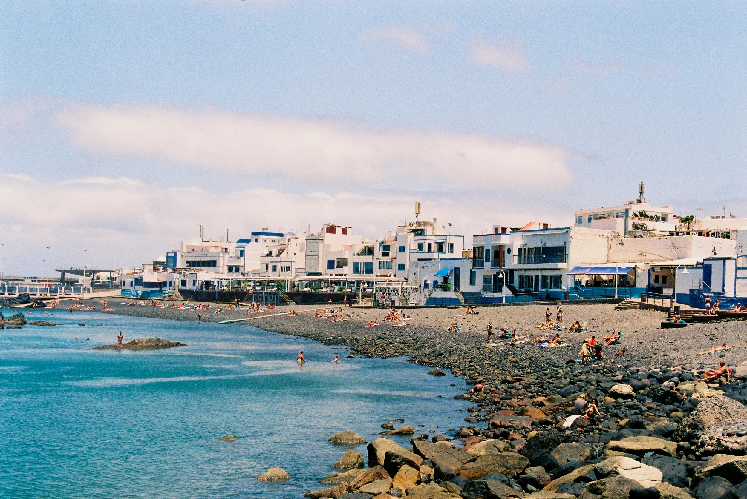A beach scene with people relaxing on the rocky shoreline and swimming in the clear blue water, with white buildings and umbrellas in the background under a partly cloudy sky.