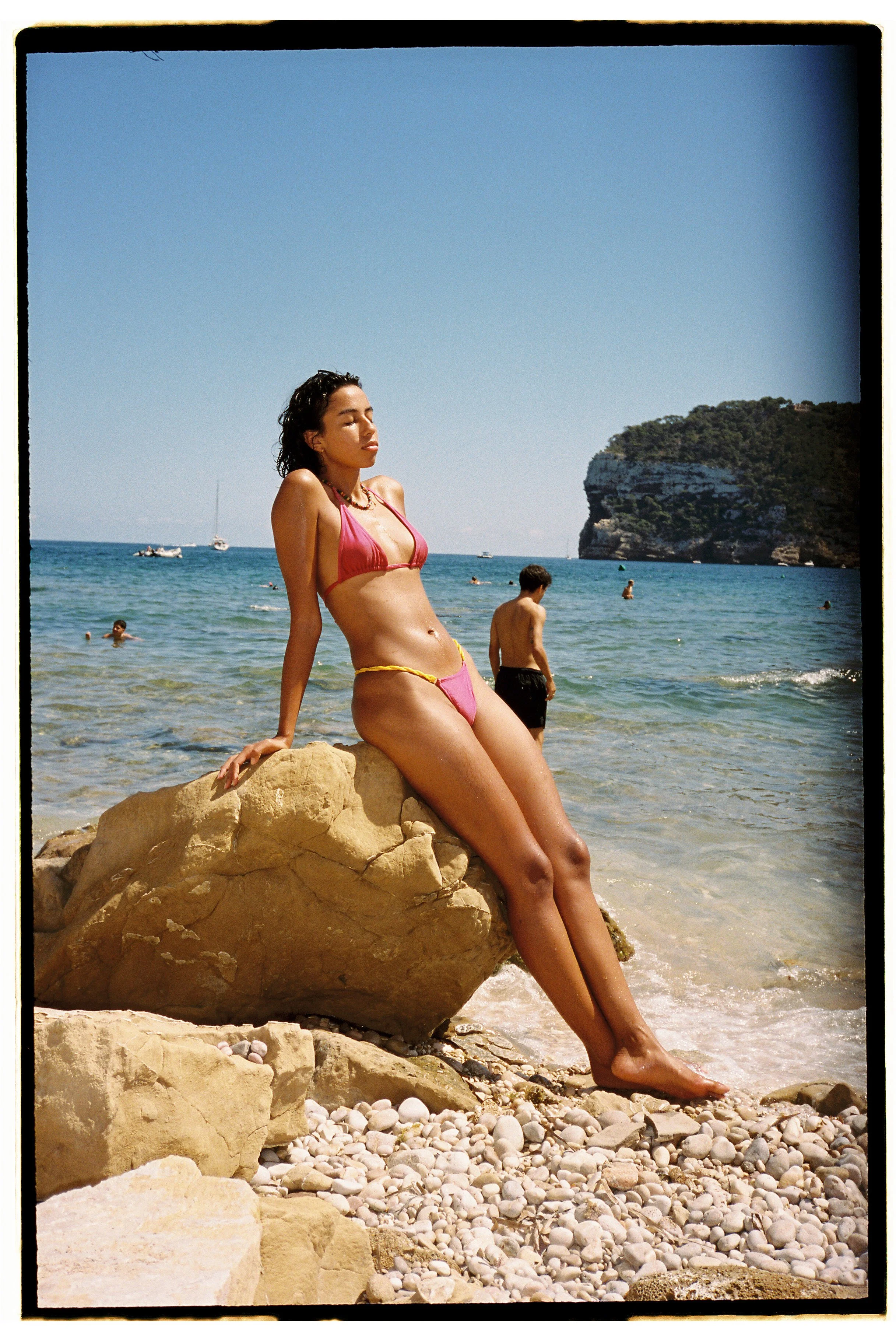 A woman in a pink bikini sitting on a large rock on a beach with pebbles, with the ocean, boats, and a rocky island in the background.