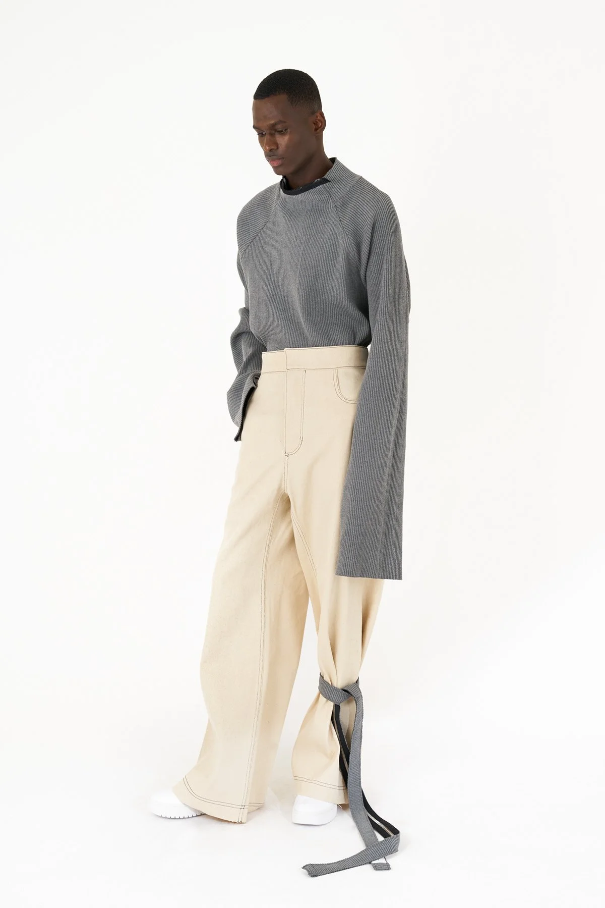 Man wearing a gray long-sleeve shirt and beige wide-leg pants with a gray strap tied around one leg, standing against a white background.