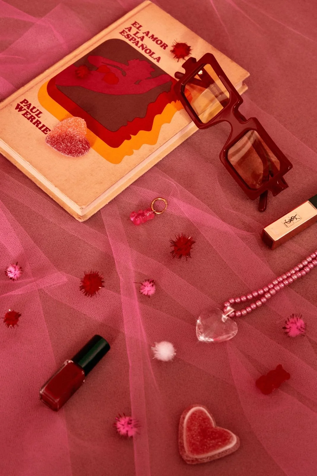 A flat lay of Valentine's Day themed items on pink fabric, including a book, glasses, nail polish, lipstick, a pink beaded bracelet with a heart charm, a small pink teddy bear keychain, and fuzzy heart and flower decorations.