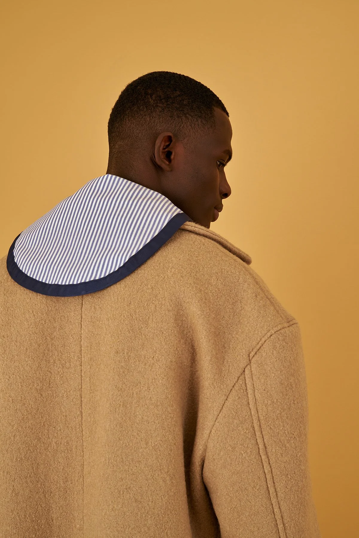 A young man in a tan coat with a striped hoodie underneath standing against a yellow background, viewed from behind in profile.