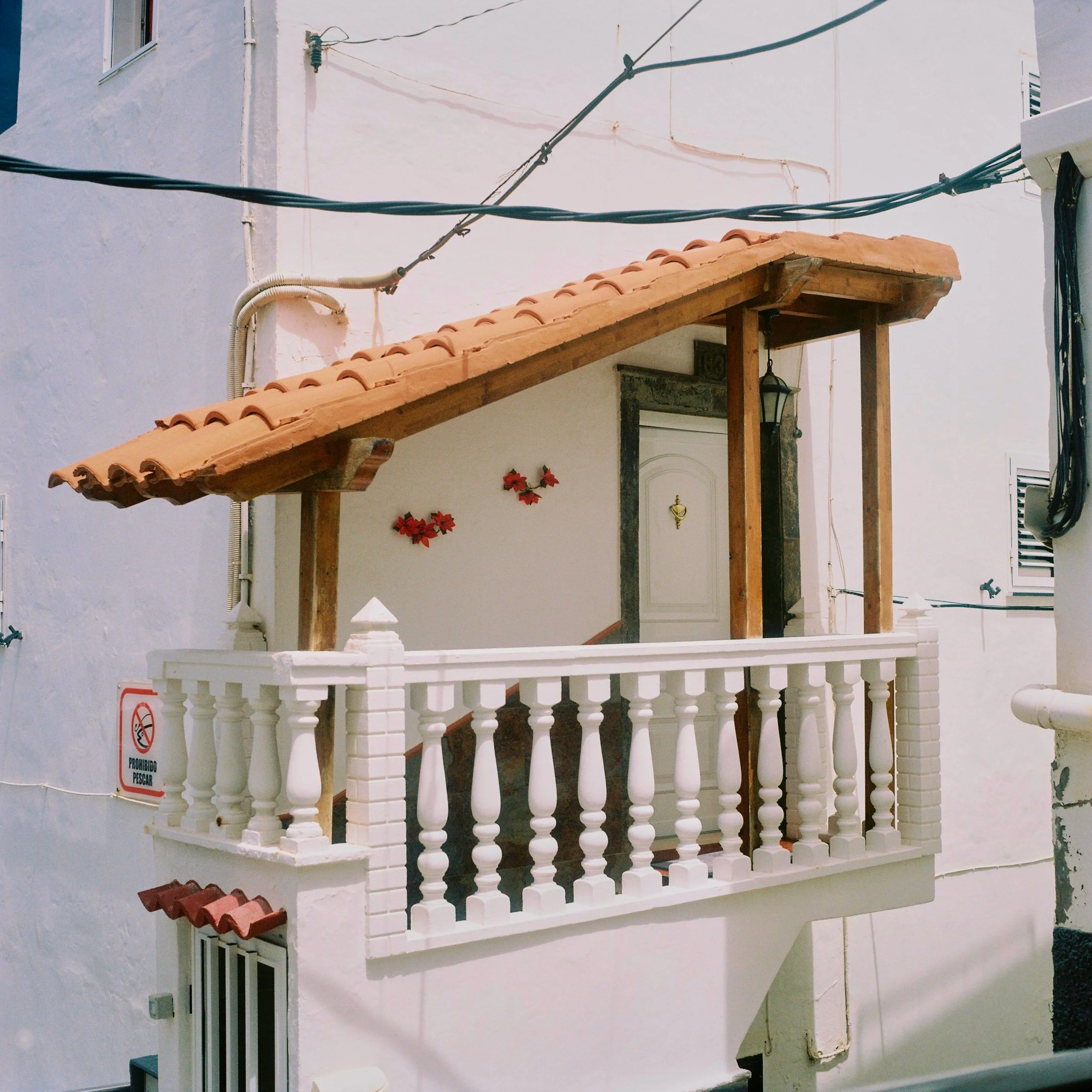A small balcony with a white brick railing, wooden posts, and a terracotta tiled roof, attached to a white building celebrating a Mediterranean or Spanish style.