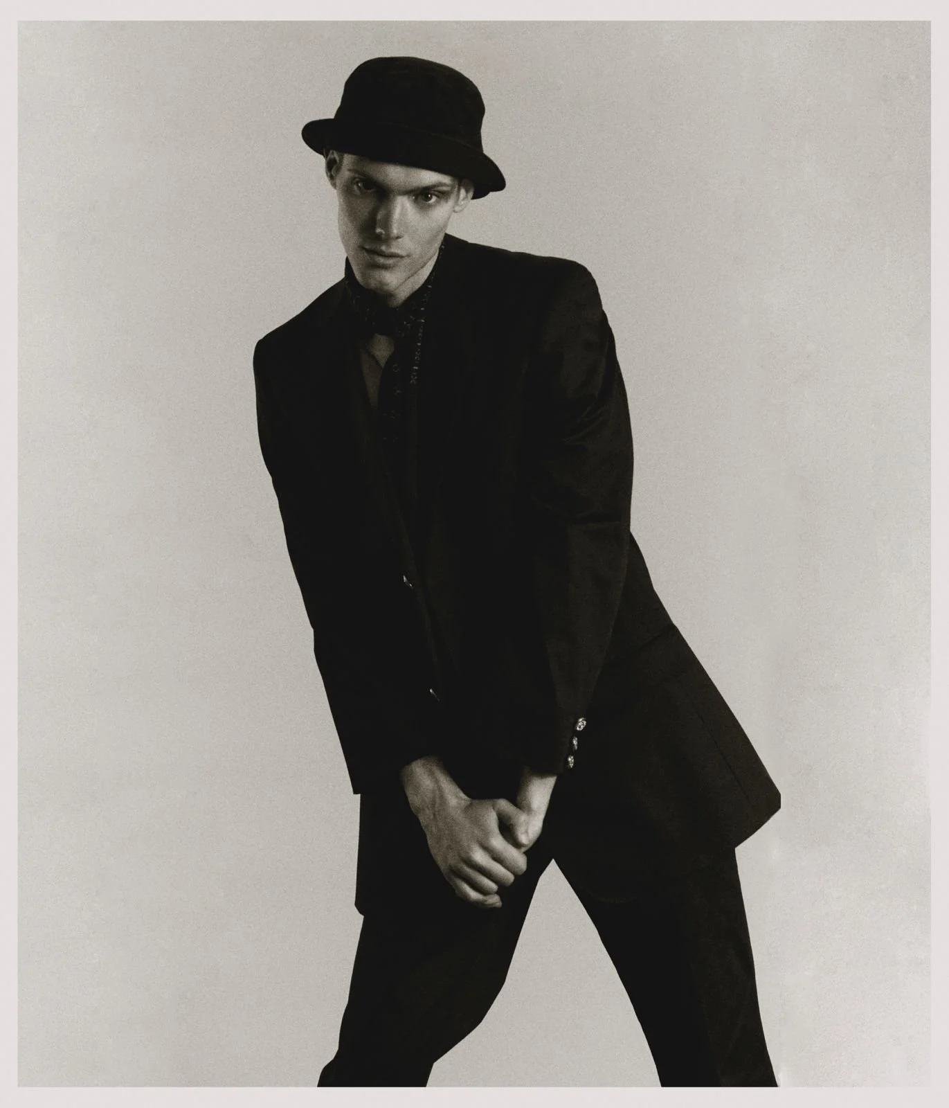 Black and white photo of a young man wearing a black hat, dark jacket, and patterned shirt, standing with hands clasped in front, looking at the camera against a plain background.