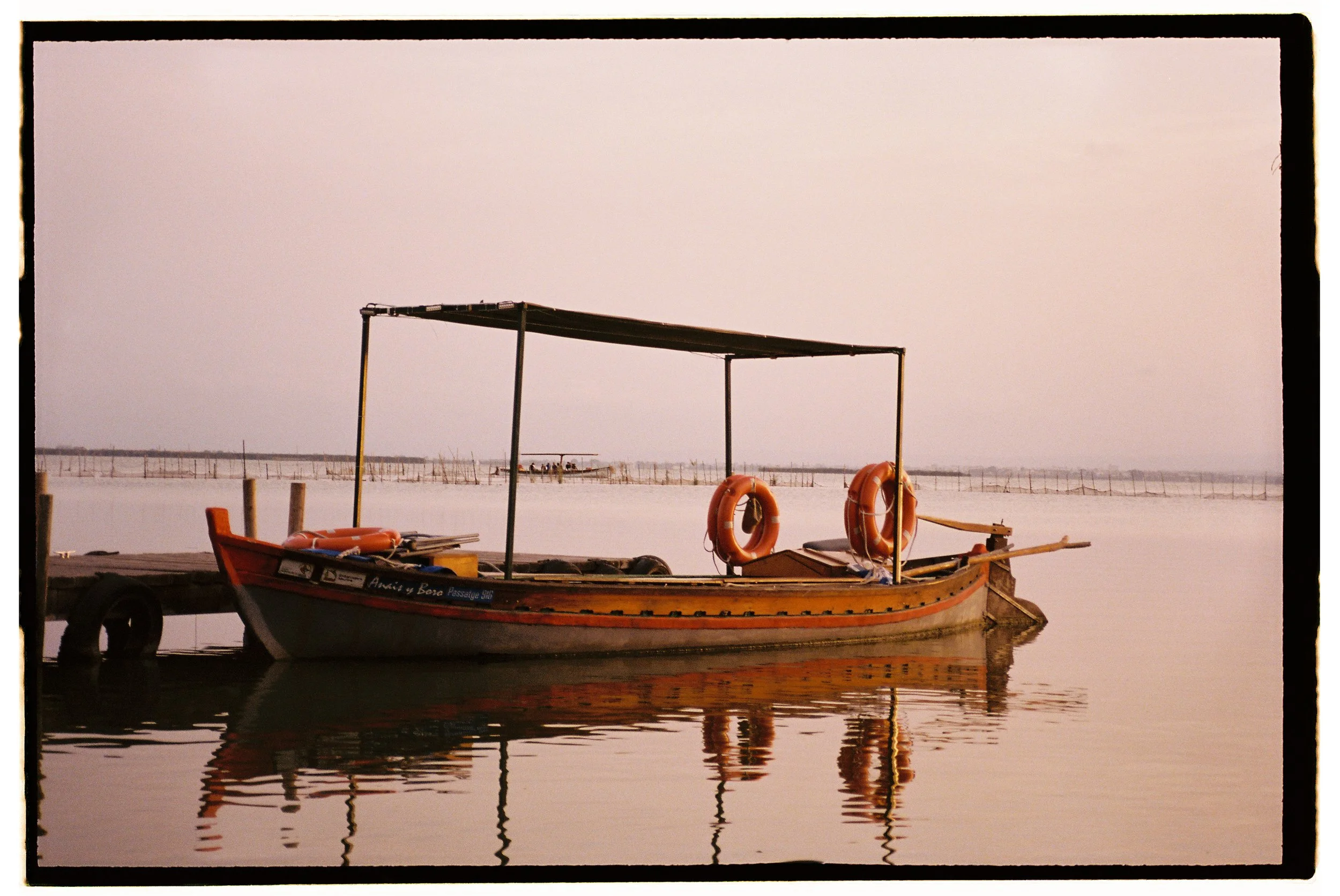 A wooden boat with orange life preservers docked along a pier, reflected in calm water near a hazy horizon at sunset.
