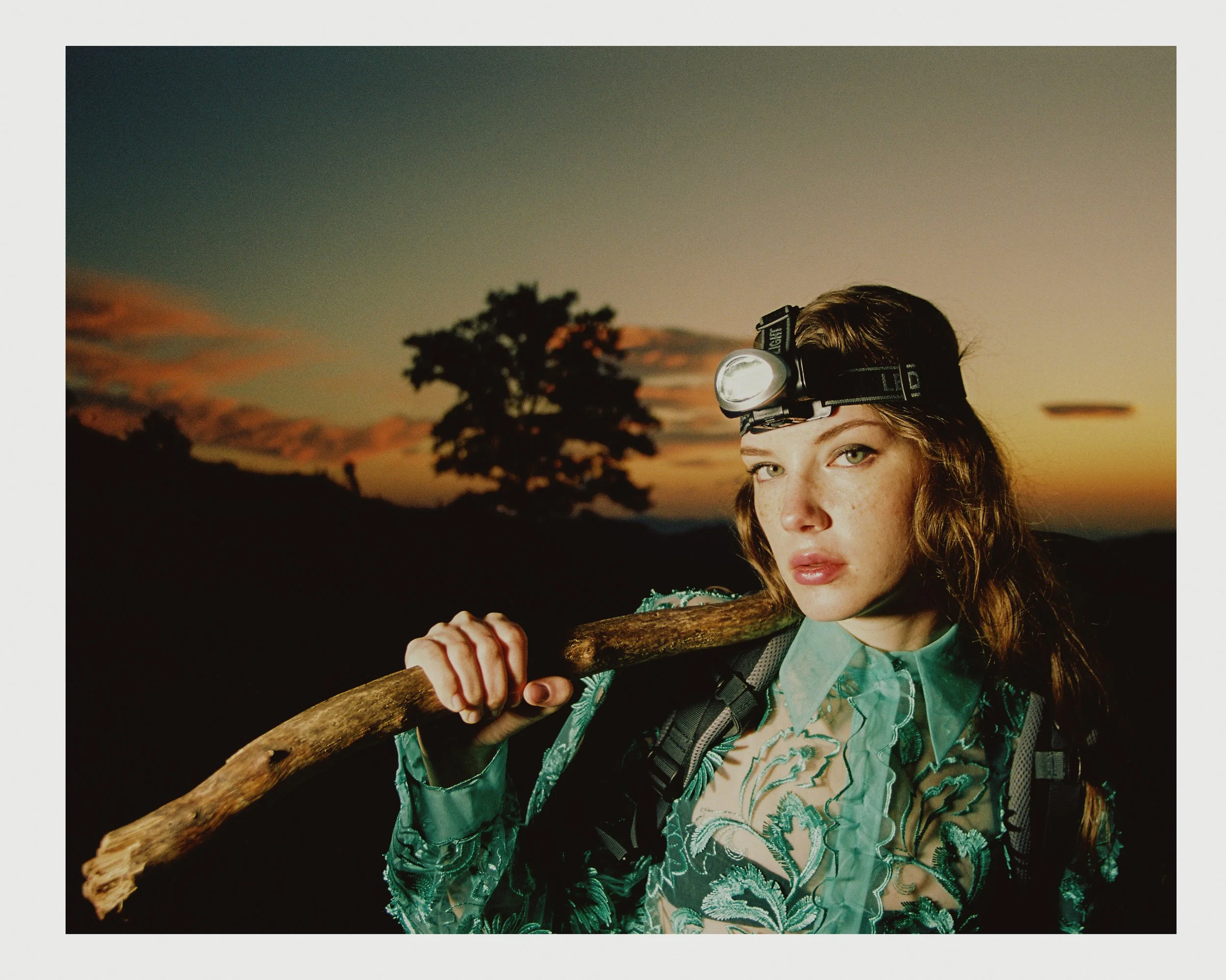 A young woman wearing a headlamp, a sheer embroidered blouse, and a backpack, carrying a wooden stick on her shoulder during dusk with silhouette trees and a sunset in the background.