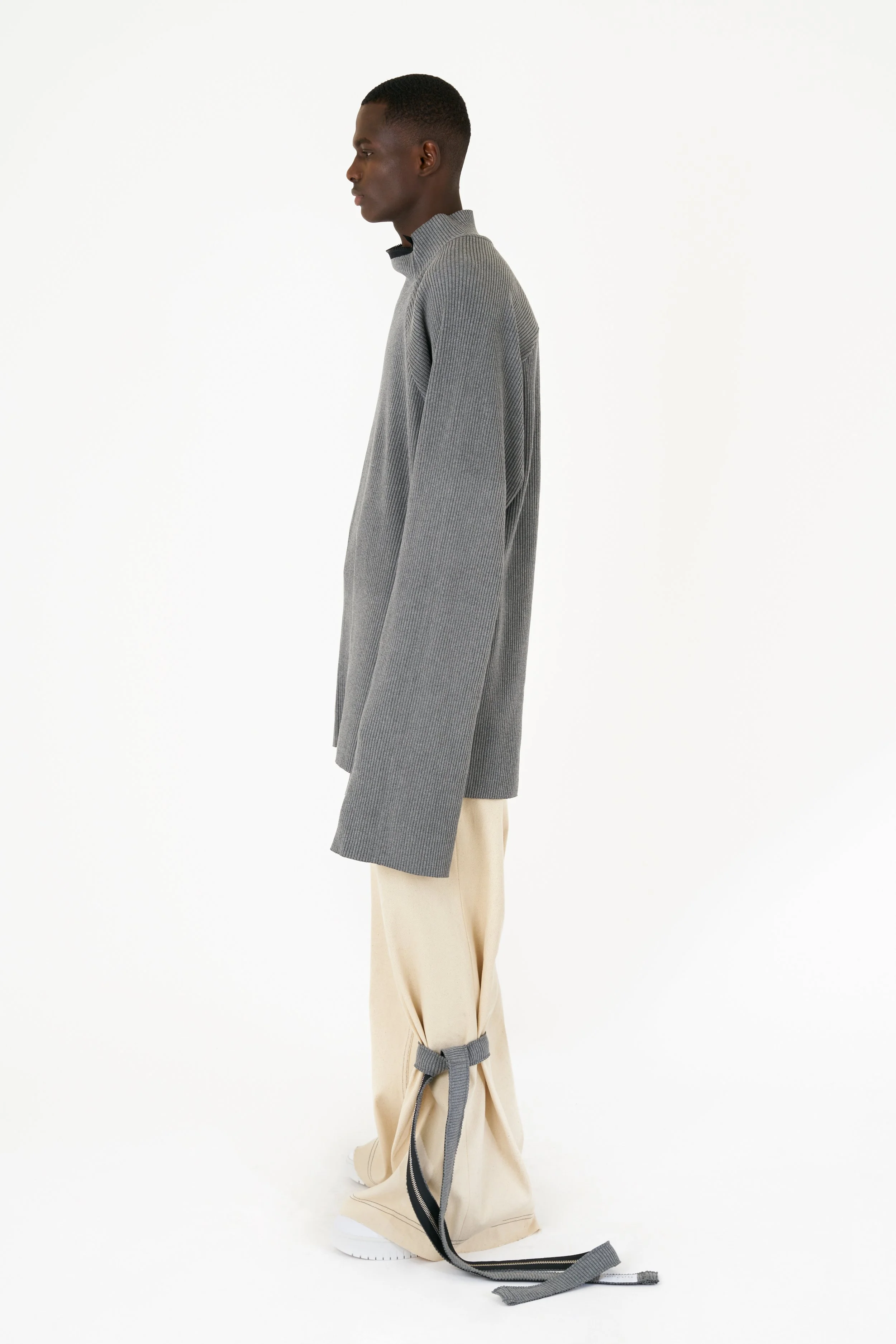 Side view of a young man in a gray high-neck sweater and beige pants with ties at the bottom, standing against a white background.