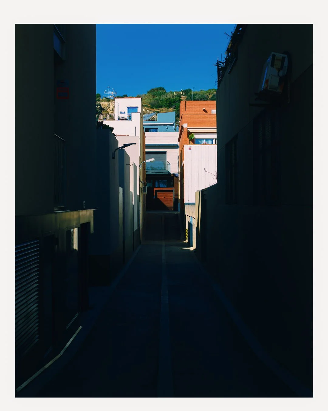 Narrow alley between modern residential buildings with shadows, leading to colorful houses under a clear blue sky.