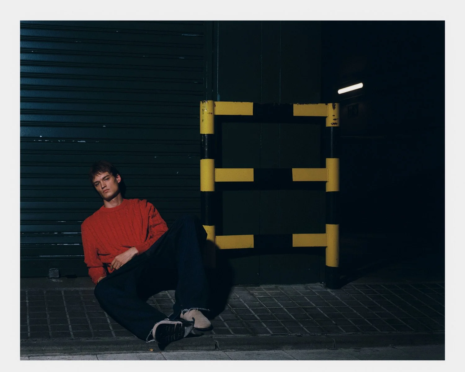 A young man in a red sweater and dark pants sitting on the sidewalk at night next to a yellow and black barrier in front of a dark, closed storefront with a metal roller shutter and a single overhead light.