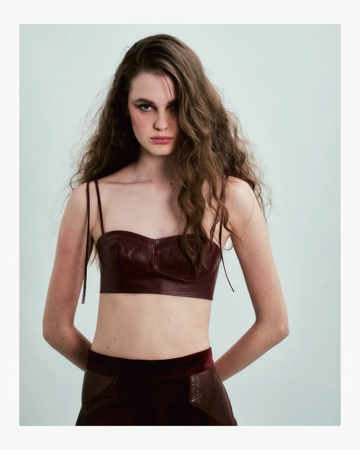 Young woman with wavy brown hair, wearing a dark red leather crop top with spaghetti straps, standing against a plain light background.