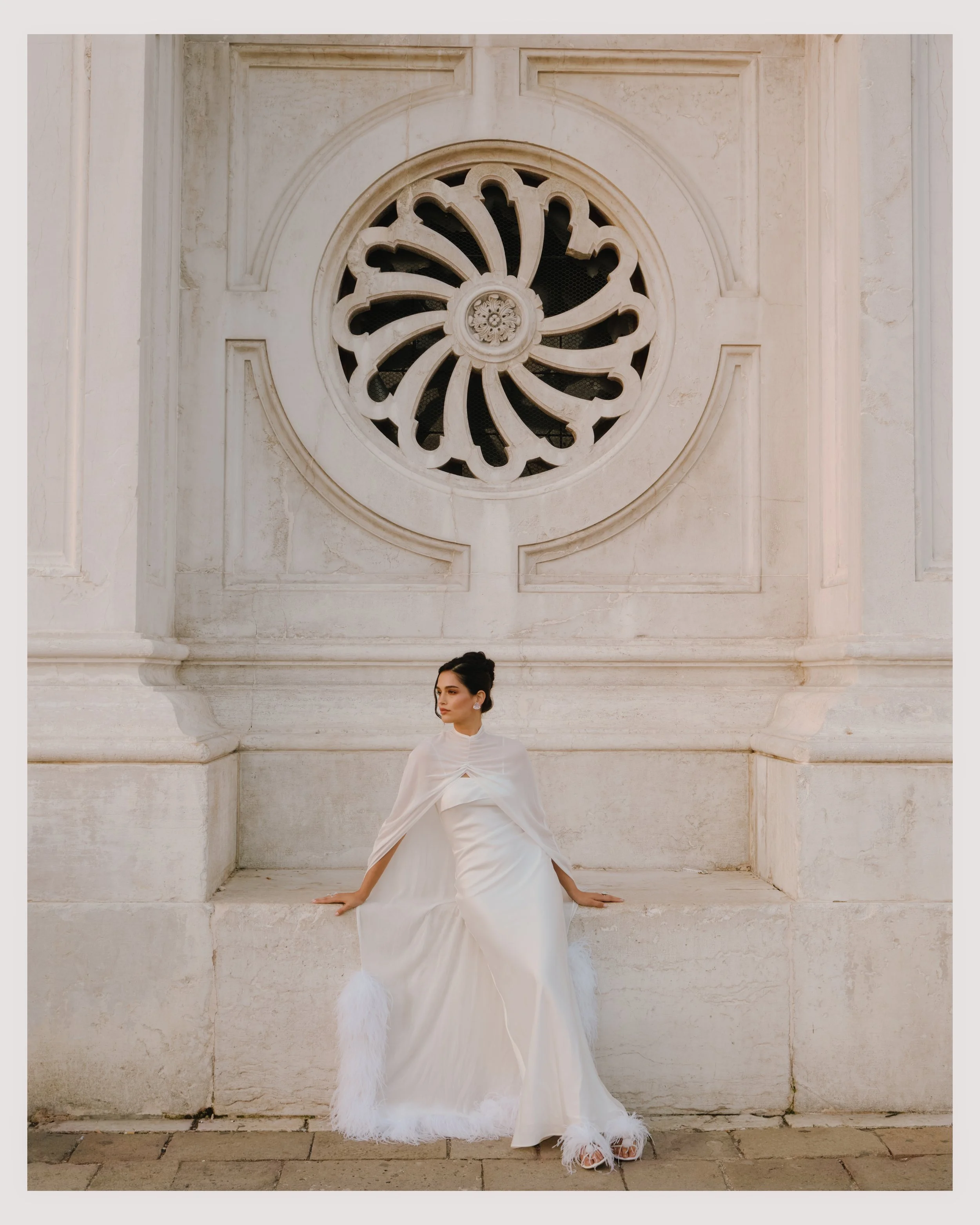A woman wearing a white dress and cape with feathered hem, sitting against a stone ledge in front of a large white wall with a decorative circular window.