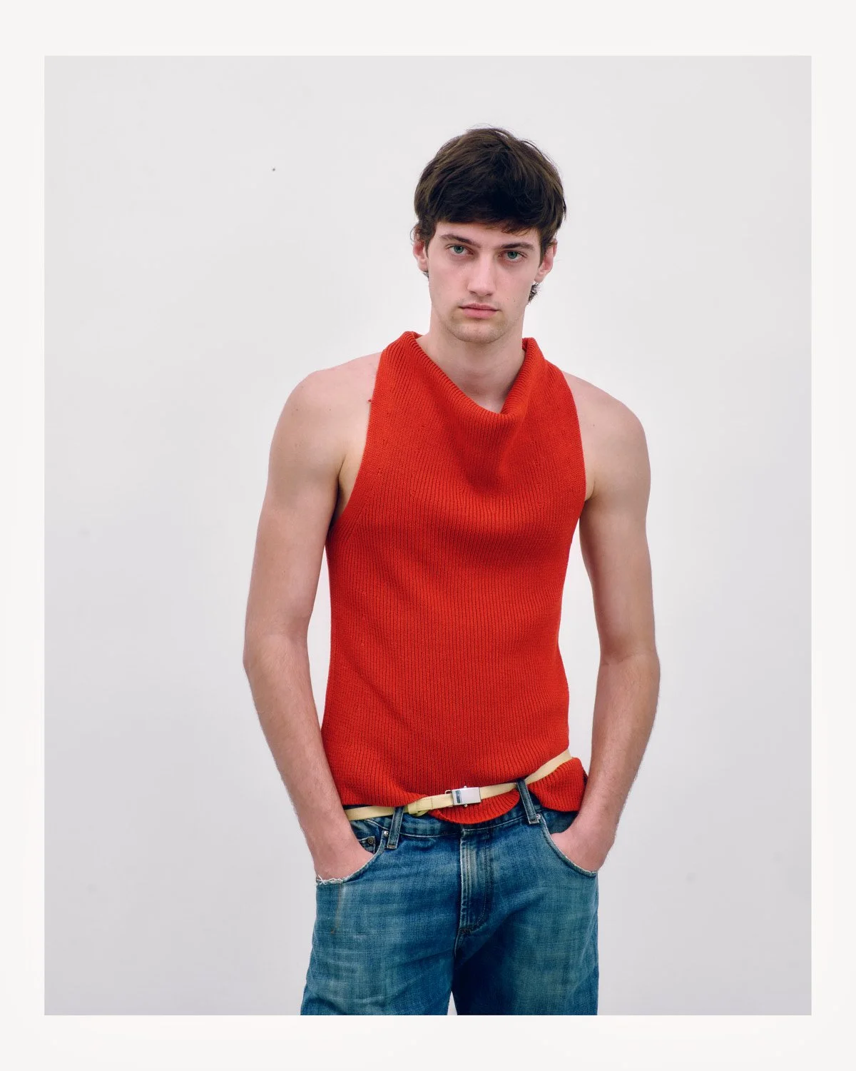 Young man with dark hair wearing a sleeveless red sweater and blue jeans against a plain white background.