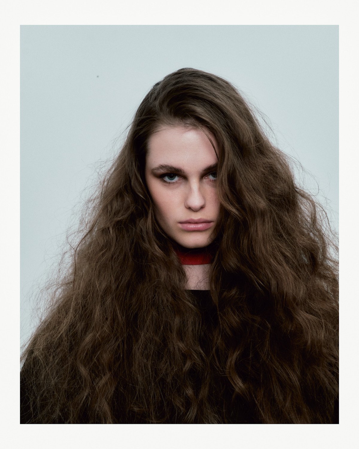 Young woman with long, voluminous, curly brown hair and piercing blue eyes, wearing a dark top and a red choker, against a plain light background.