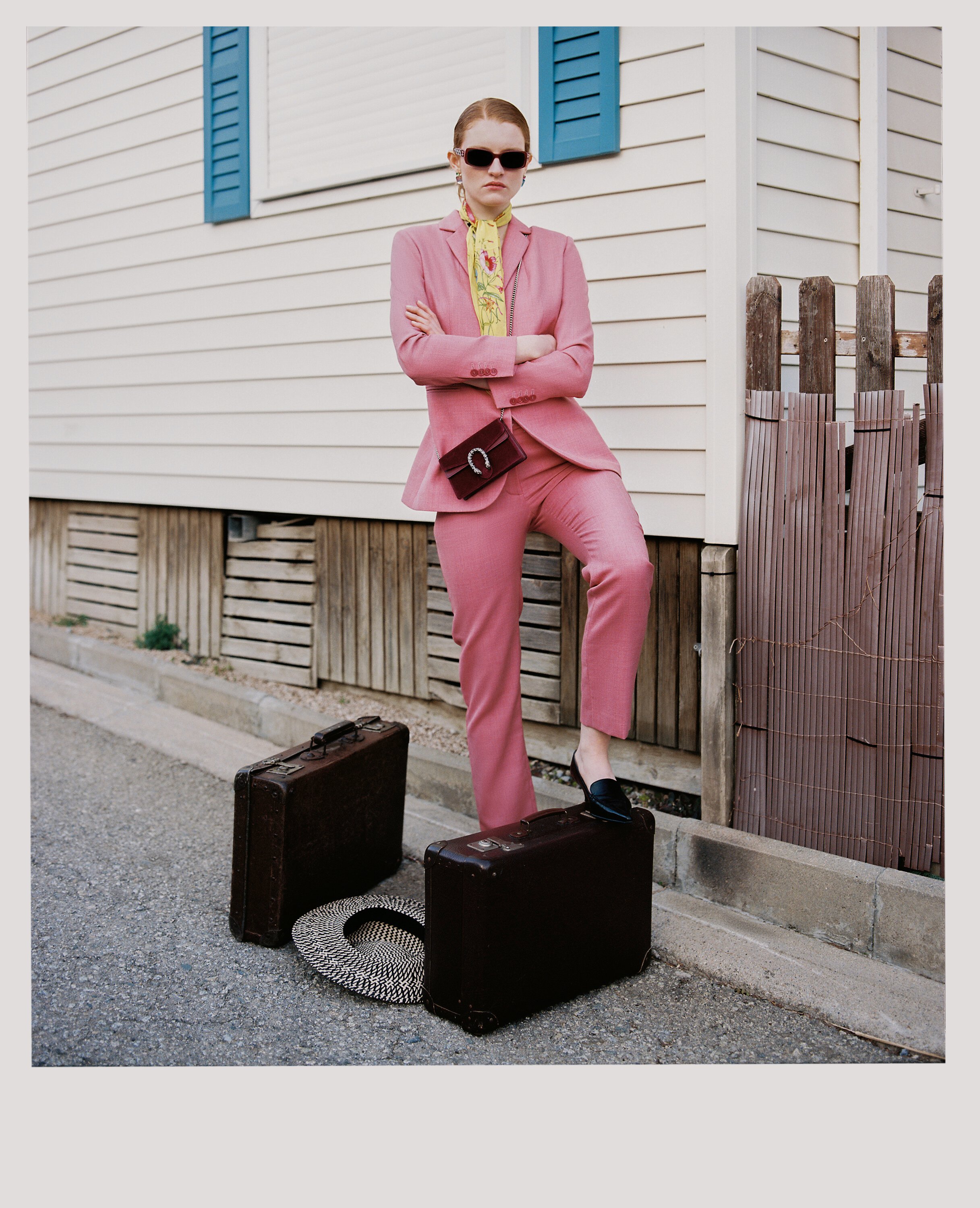 A woman dressed in a pink suit with sunglasses, standing with arms crossed on a sidewalk in front of a house with white siding and blue shutters. She has a yellow scarf, a small handbag, and shoes on a suitcase.