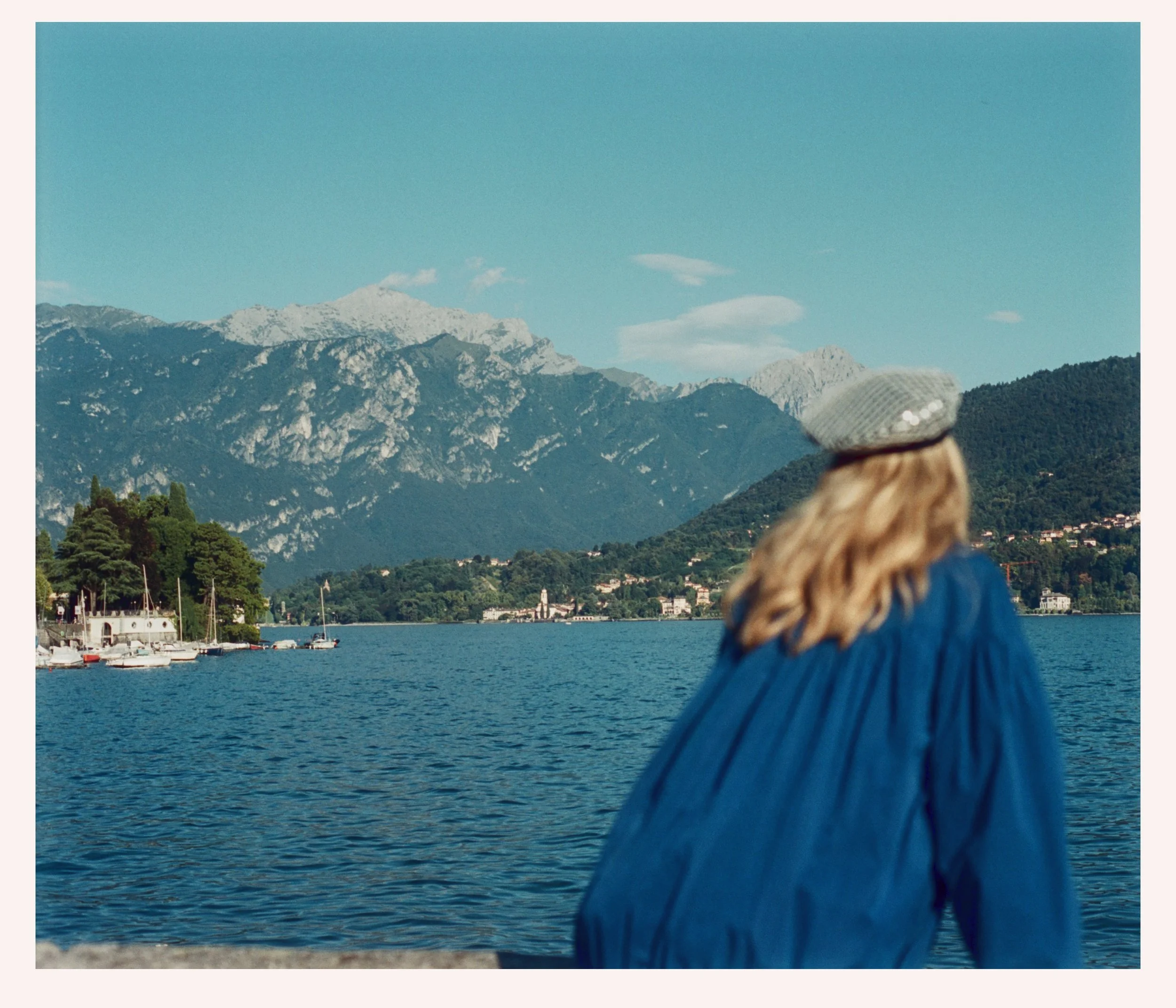 A woman with blonde hair wearing a gray beret and a blue jacket, overlooking a lake with mountains in the background.