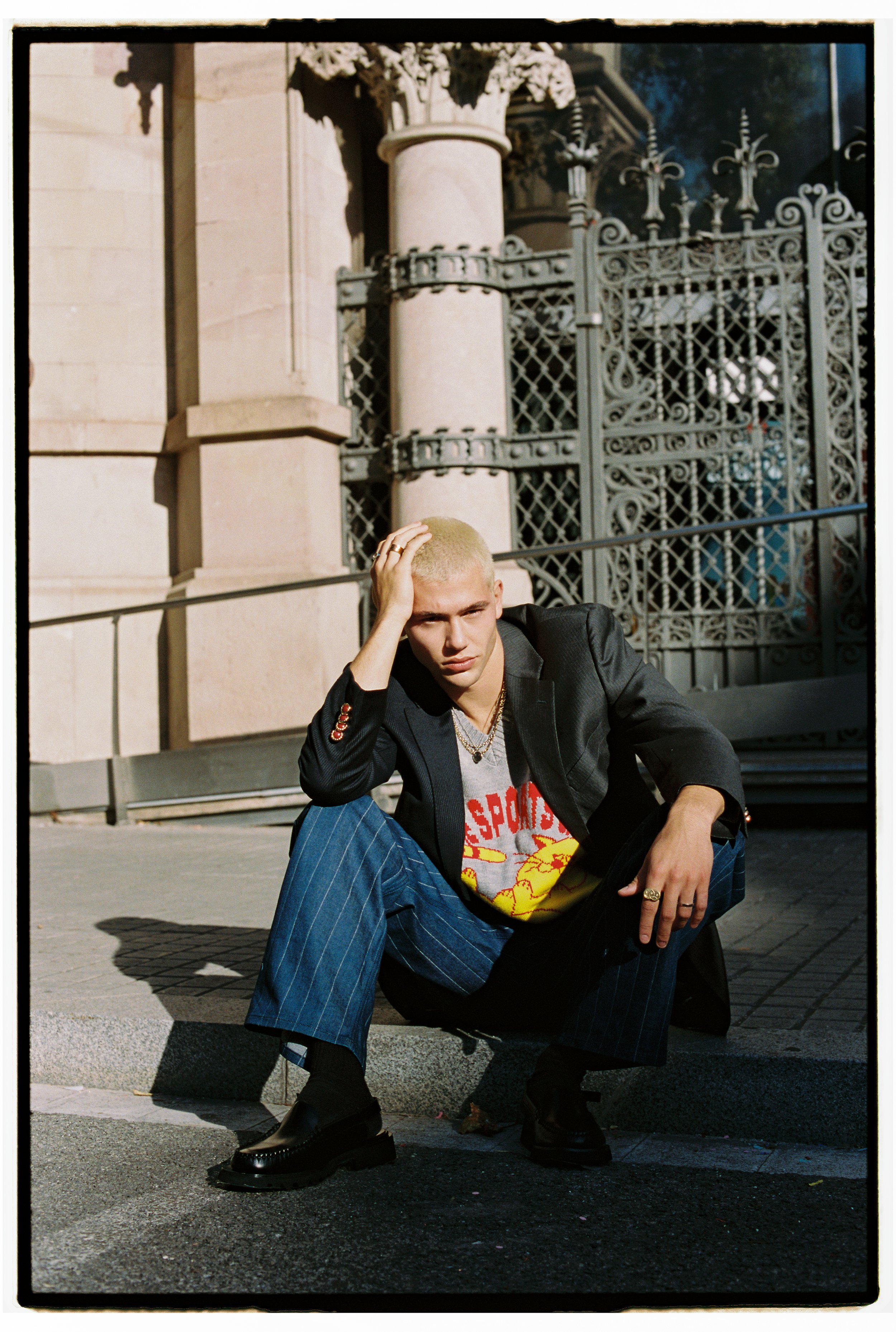 Young man with bleached blonde hair crouches on sidewalk, wearing a black blazer over a graphic t-shirt, plaid pants, and chunky black shoes, with a distressed expression, in front of ornate iron gate and classical building.