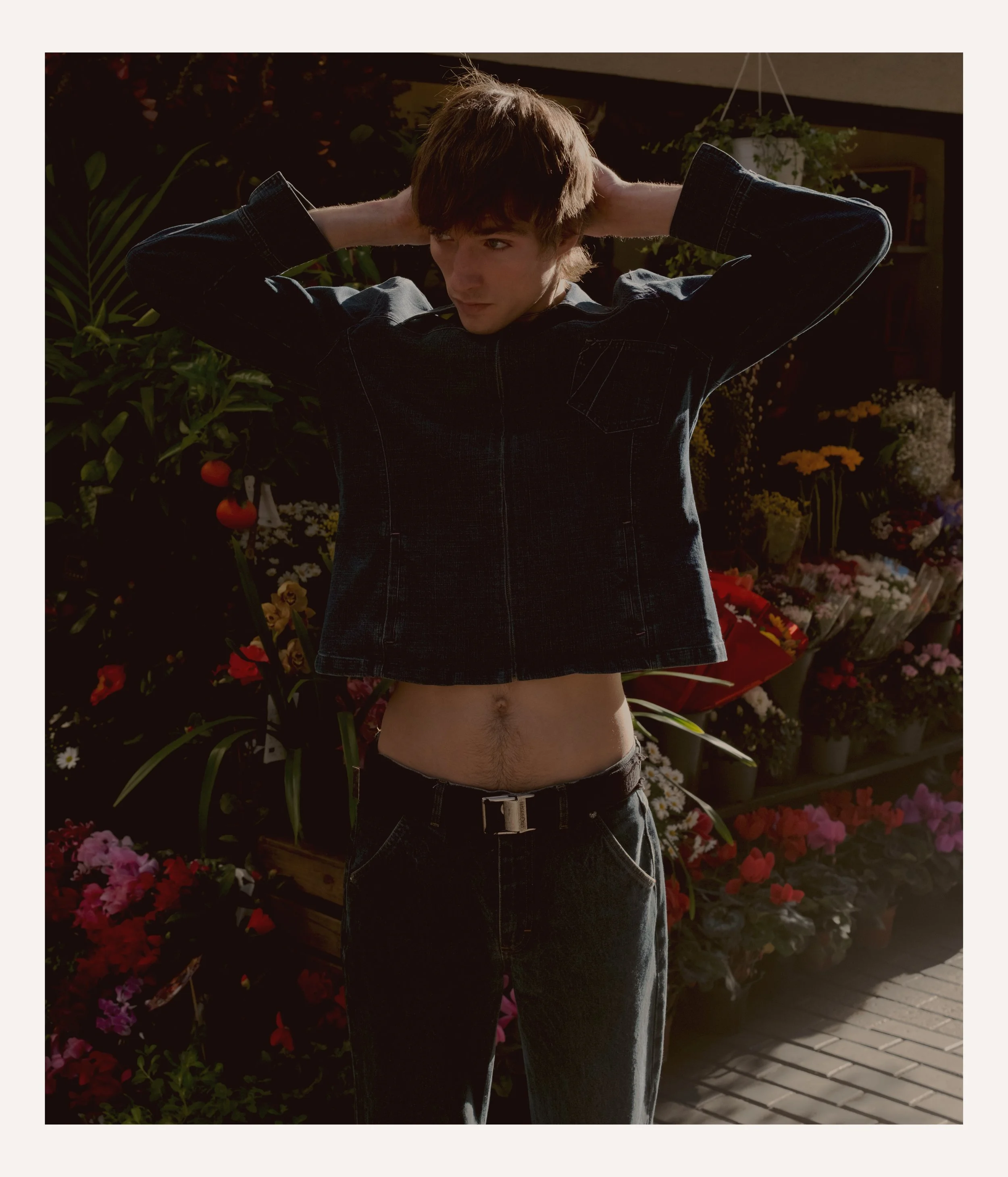 Young man standing outdoors among flowers, lifting his dark denim jacket to reveal his waist and exposed skin.