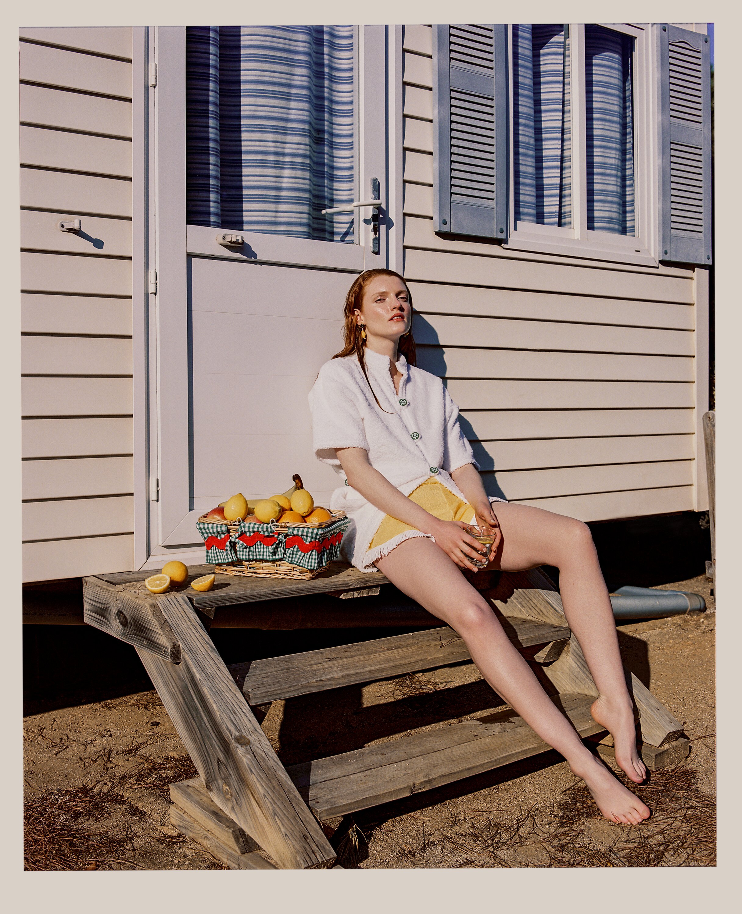 A woman sitting barefoot on wooden steps outside a house, holding a drink, with a basket of lemons and oranges beside her.