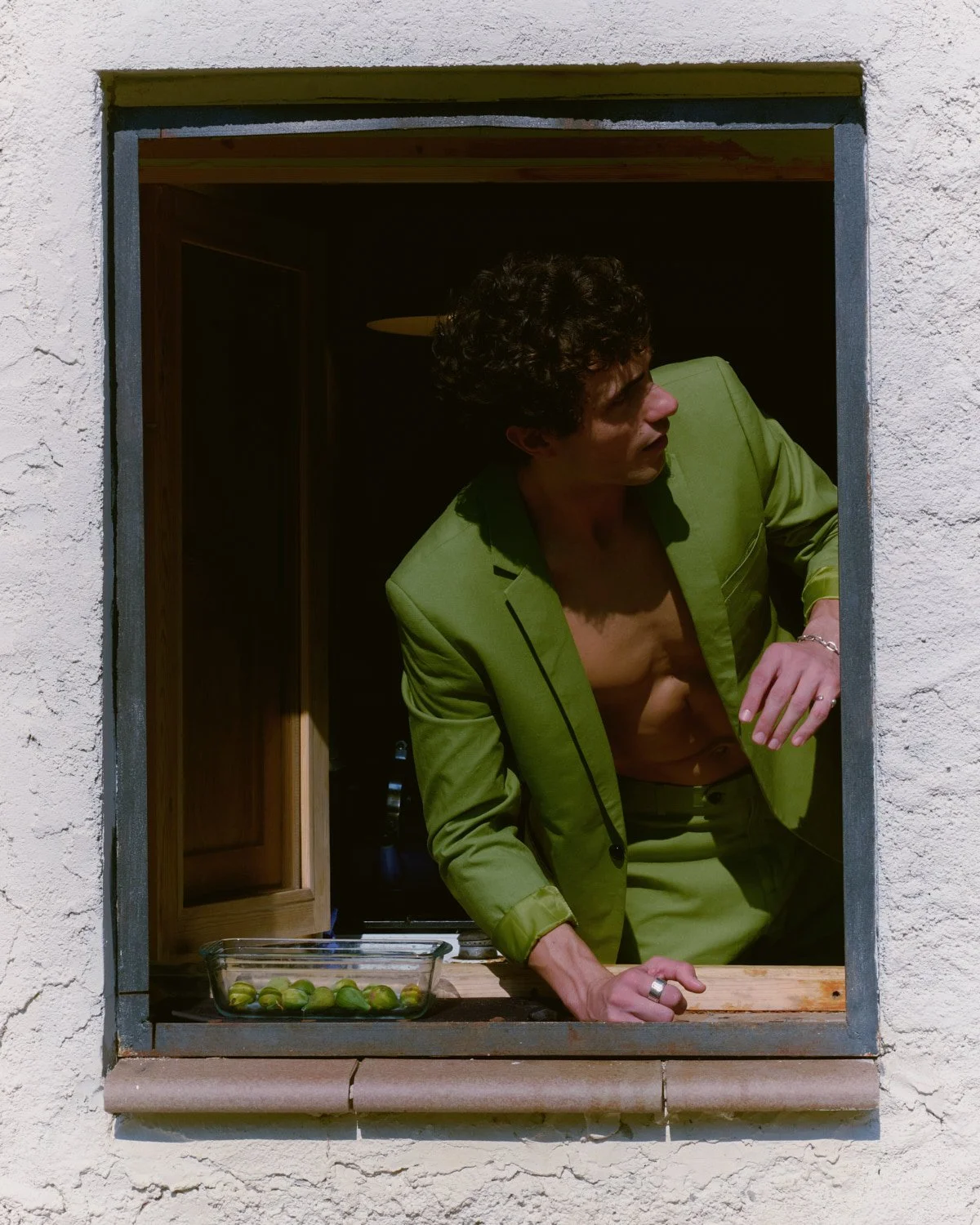 A man in a green suit leaning out of a window with a bowl of green fruits on the window ledge.