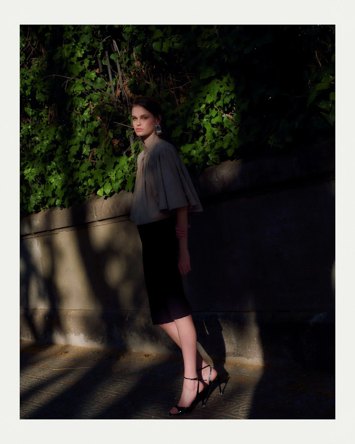 A woman with dark hair in a grey cape top and black skirt, wearing black high heels, standing outdoors against a background of green leafy bushes and a stone wall, with shadows cast on the ground.