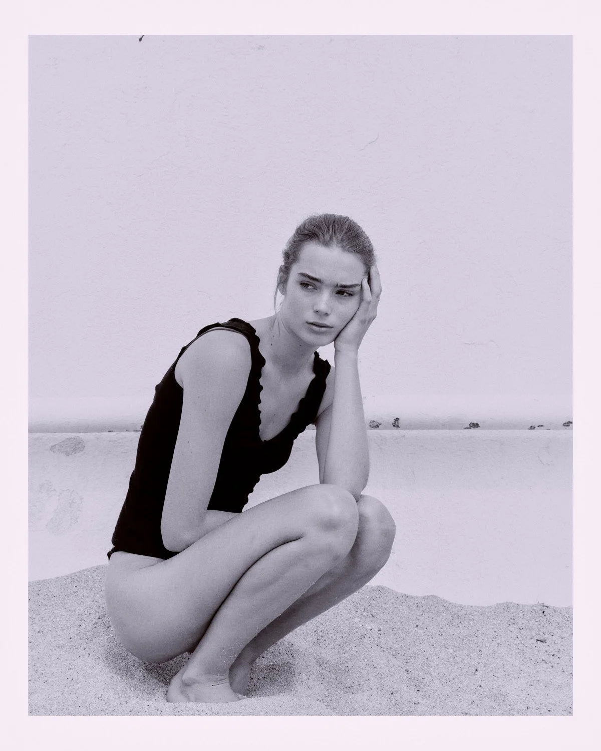 Black and white photo of a woman crouching on a beach, wearing a black sleeveless top, with her hair tied back, holding her head with her right hand, and looking to the side with a thoughtful expression.