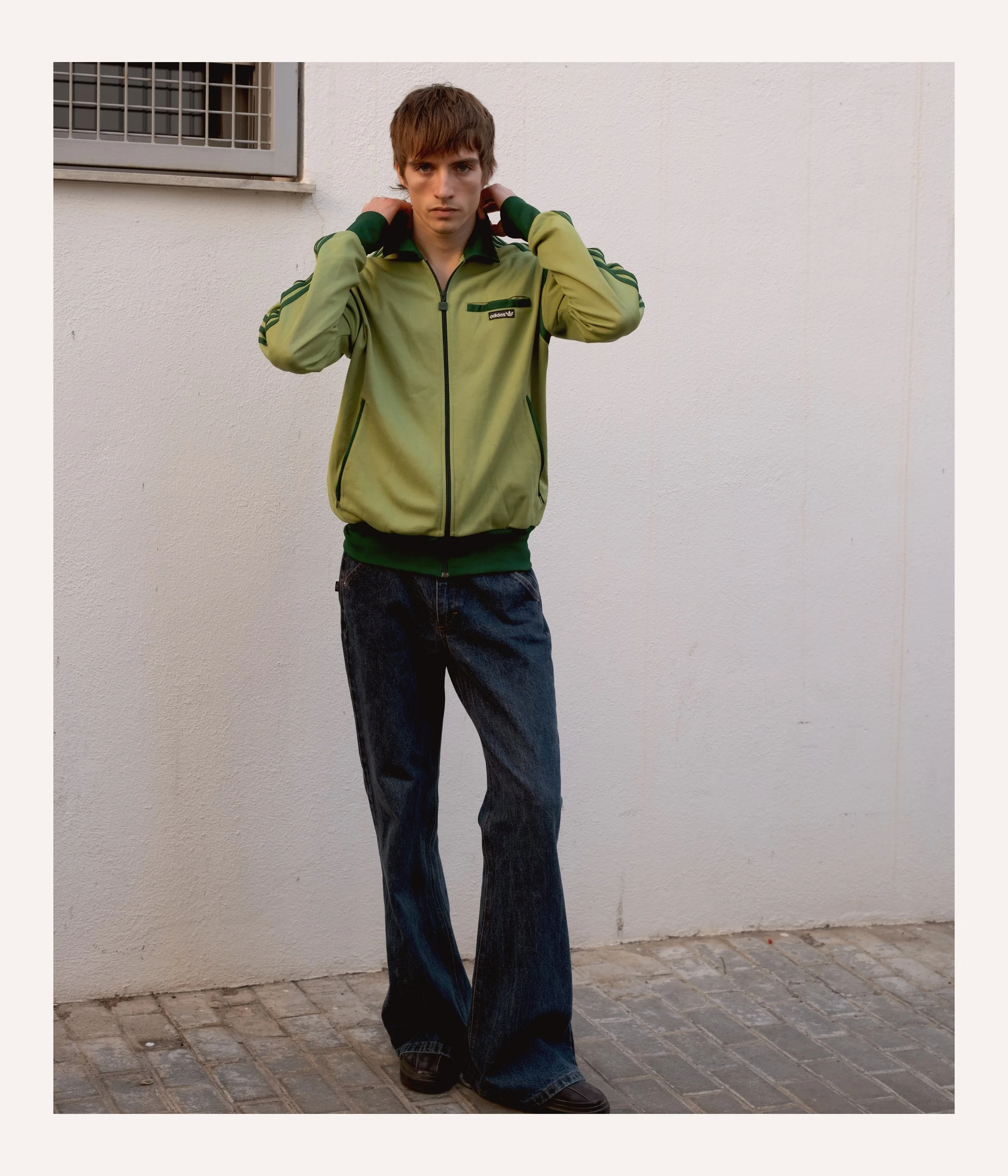 Young man standing outdoors against a plain wall, wearing a green jacket with black accents and dark blue jeans.