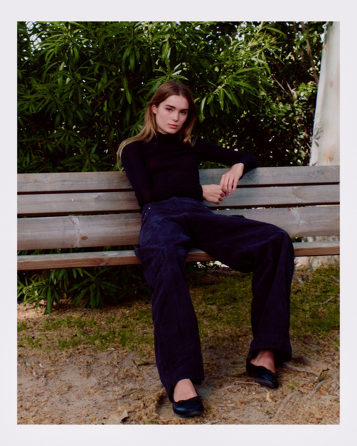 A young woman with long light brown hair, wearing a black turtleneck and dark pants, sitting on a wooden park bench with a background of green leafy bushes.