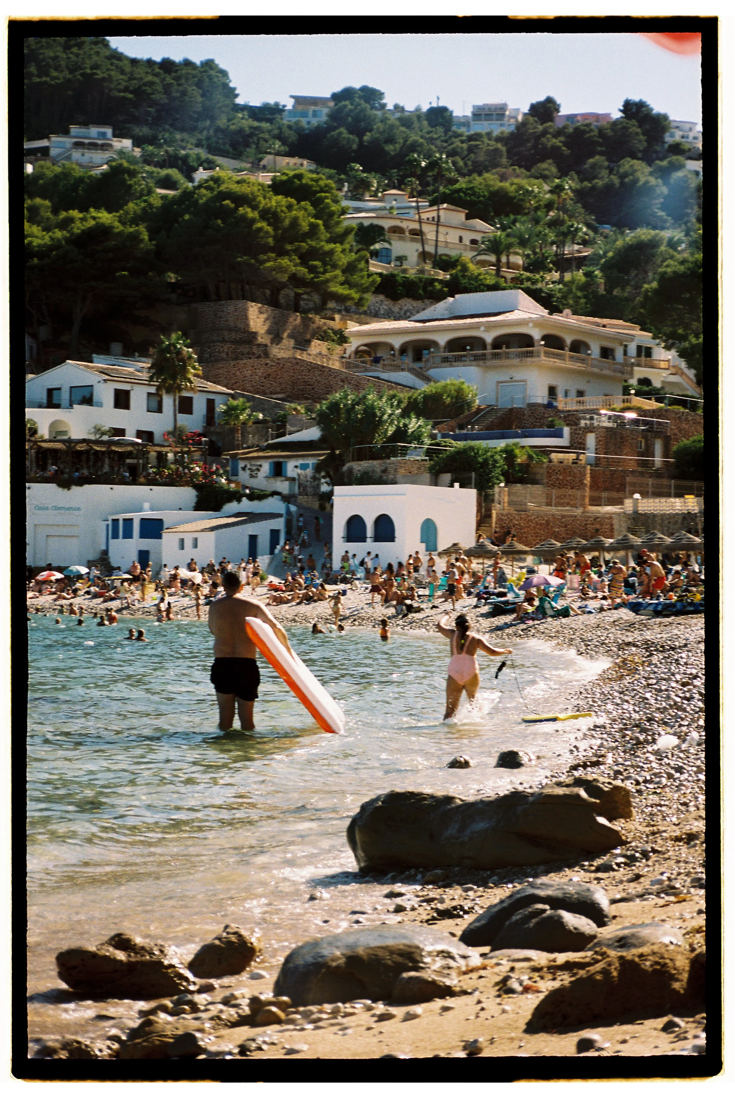 Beach scene with people swimming and relaxing, houses and greenery on hillside in the background
