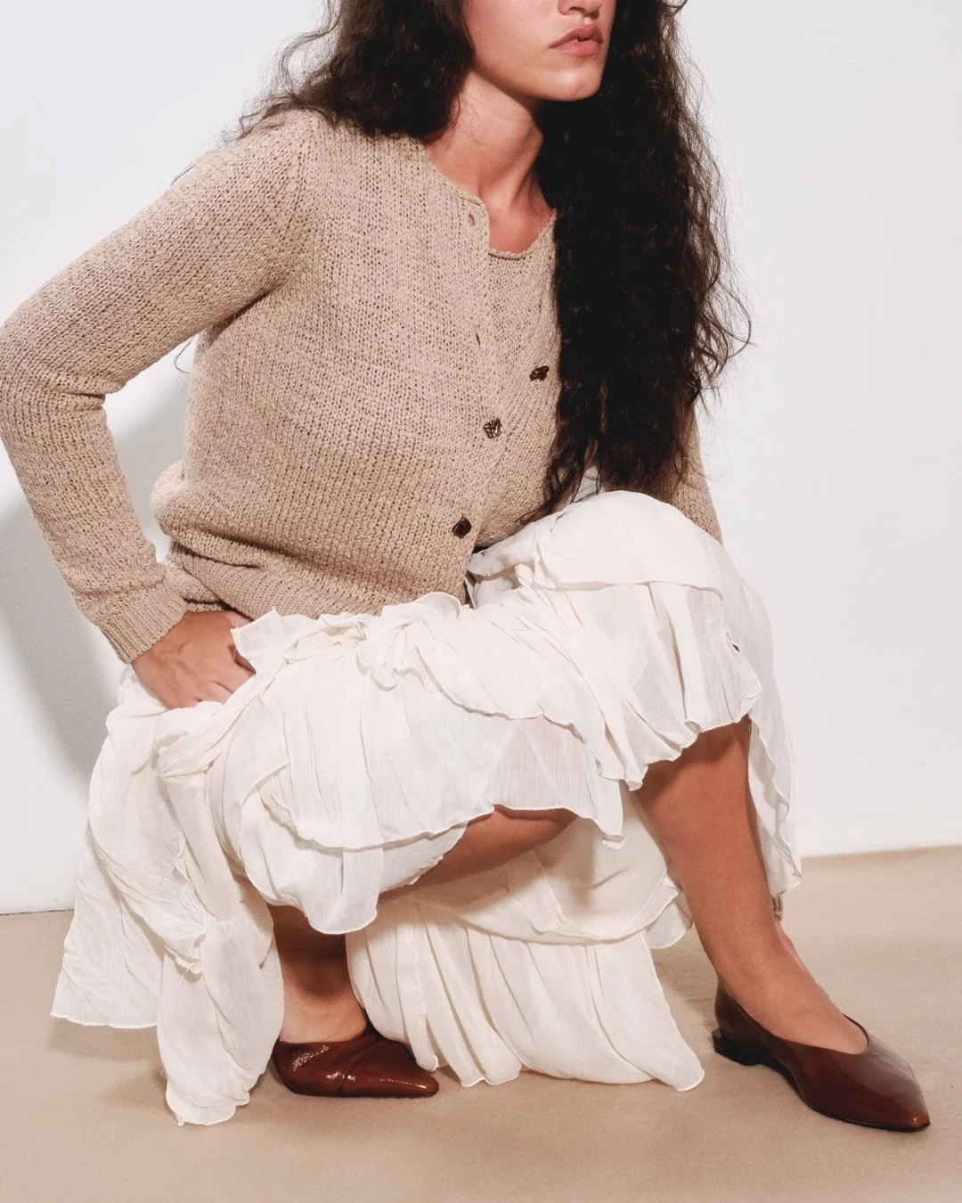 A woman with long curly hair wearing a beige cardigan and a white ruffled skirt, sitting with one leg bent and wearing brown heels.