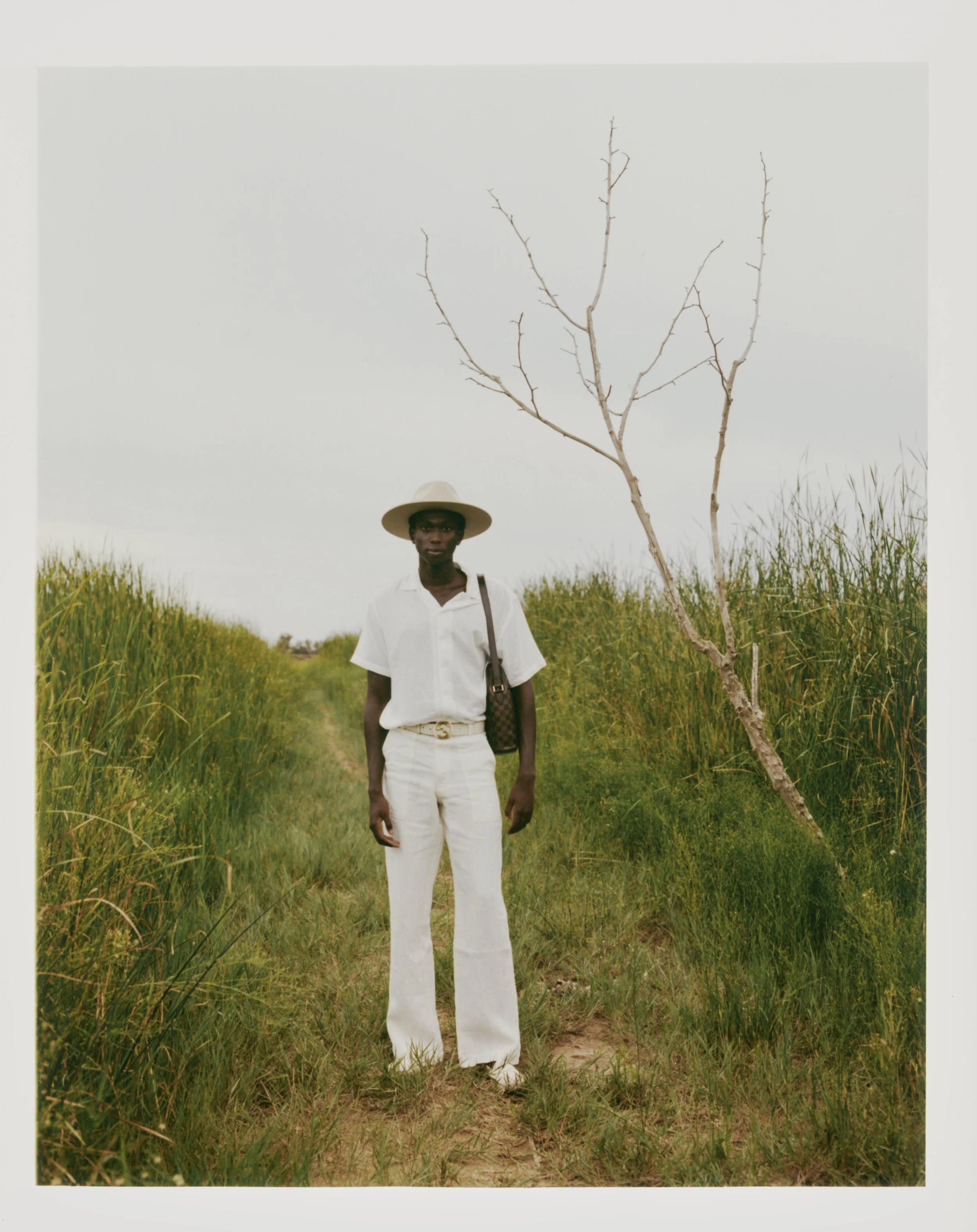 A man wearing a white short-sleeve shirt, white pants, a wide-brimmed hat, and holding a patterned bag stands on a grassy path with tall grass and a leafless tree nearby, under an overcast sky.