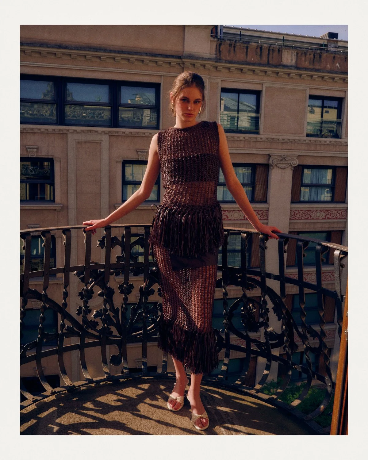 Woman in a sheer, textured dress with fringes, standing on a balcony with decorative railing, in front of a building with multiple windows.