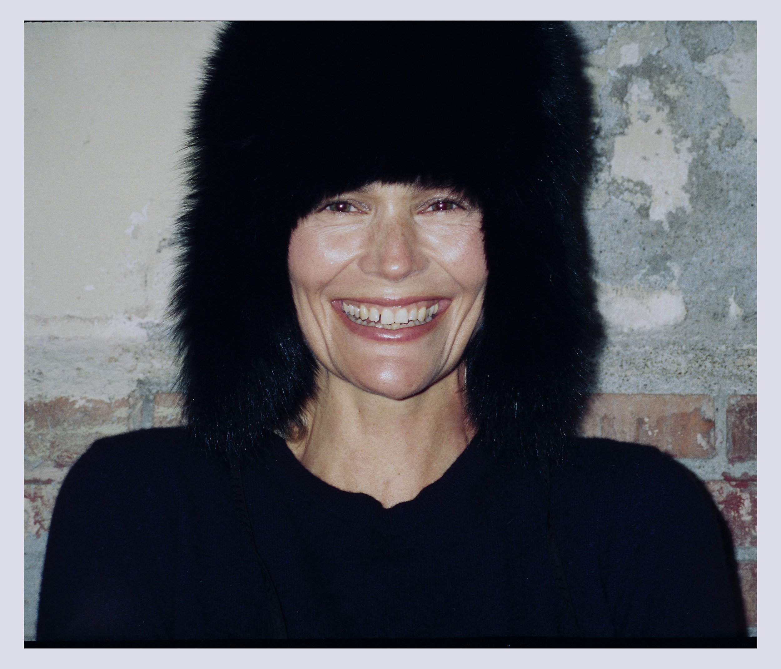 A woman smiling wearing a black fur hat and black shirt, standing in front of a brick and concrete wall.