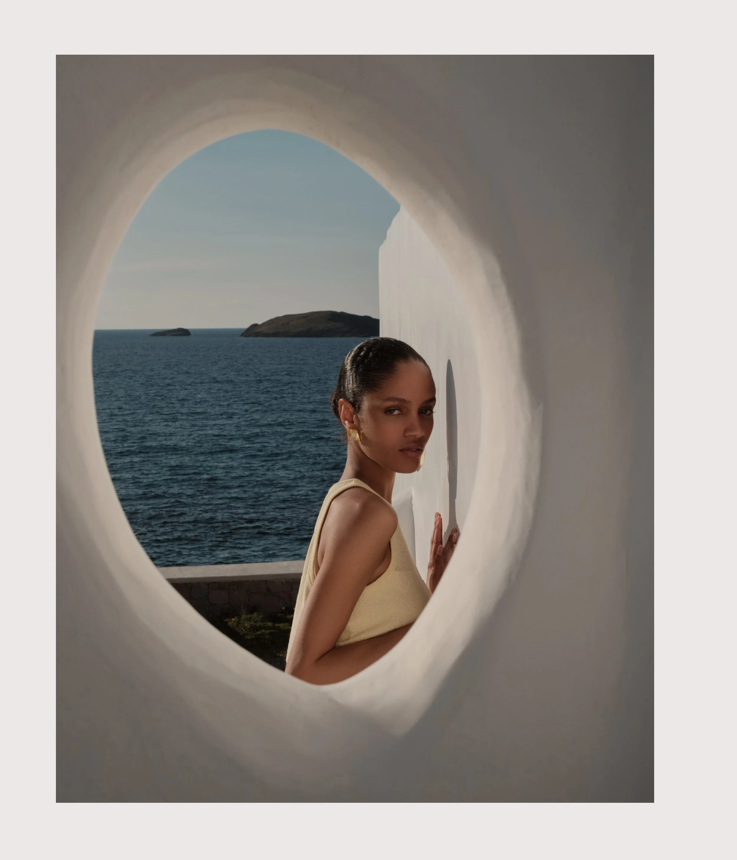 A woman with dark hair and tan skin looking through a circular window, with a view of the ocean and rocks in the background.
