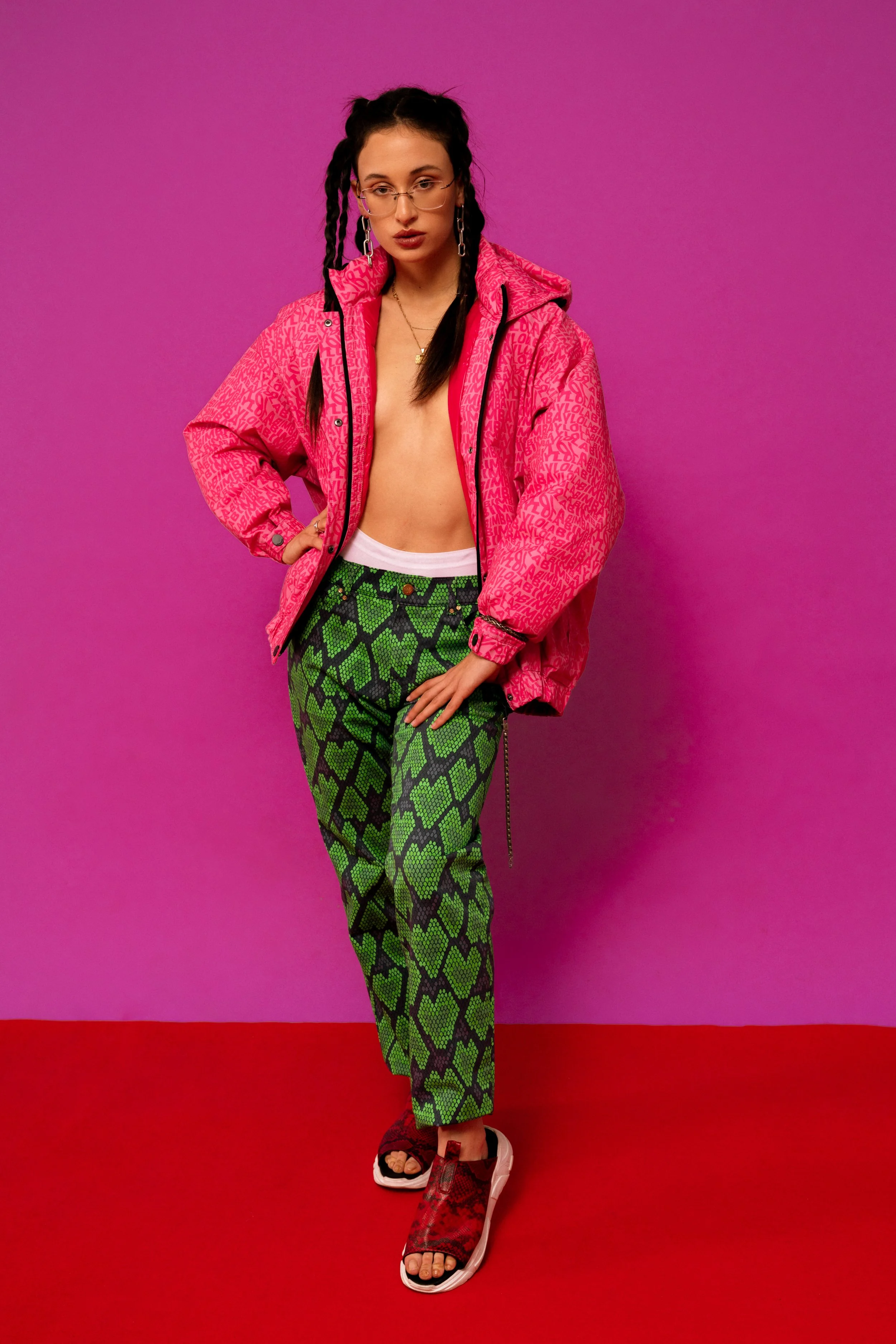 A woman in a pink patterned jacket, green snake print pants, and red sandals stands against a pink background.