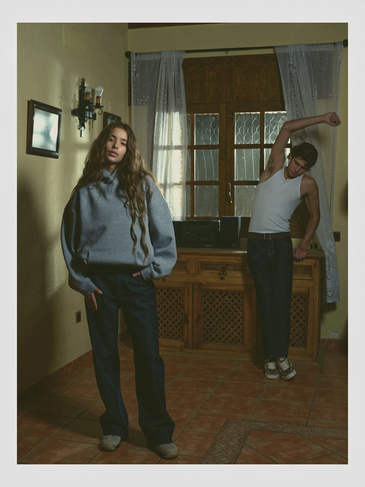A young woman with long wavy hair wearing a grey hoodie and dark jeans standing near a yellow wall, and a young man with dark hair wearing a white tank top and dark jeans stretching near a window in a room with wooden furniture and patterned curtains